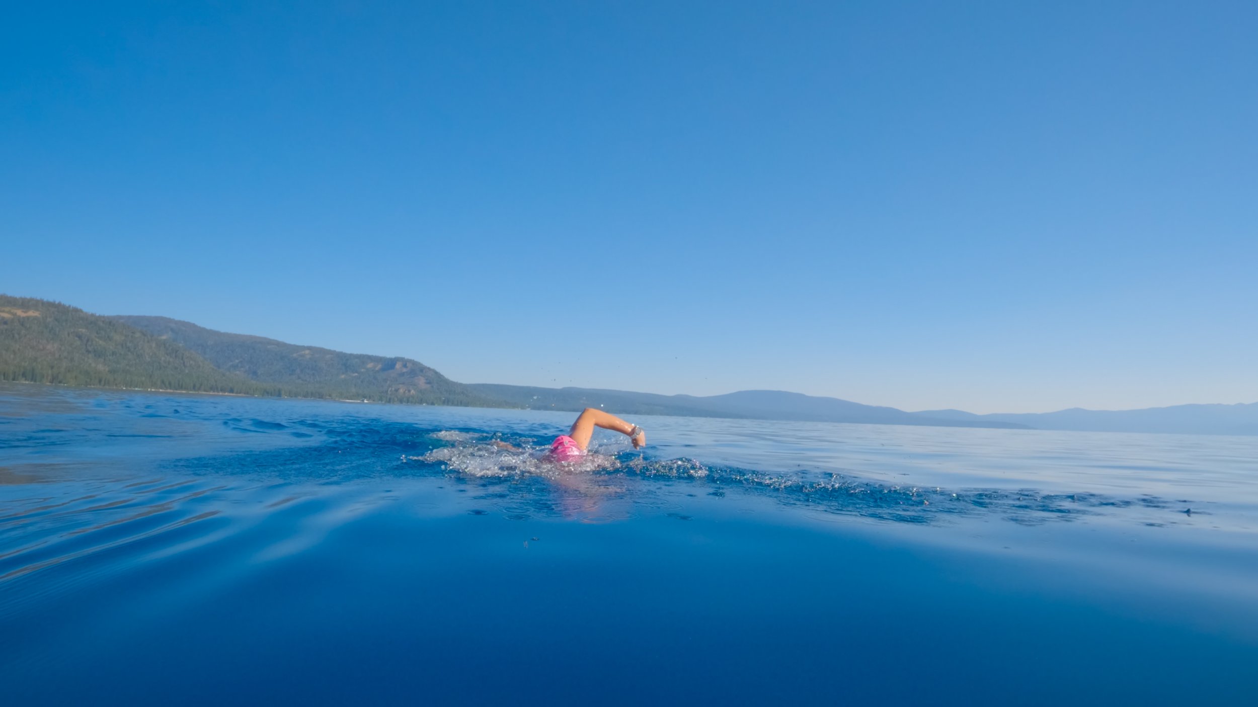 Person swimming in a large body of water with mountains in the background and a clear blue sky.