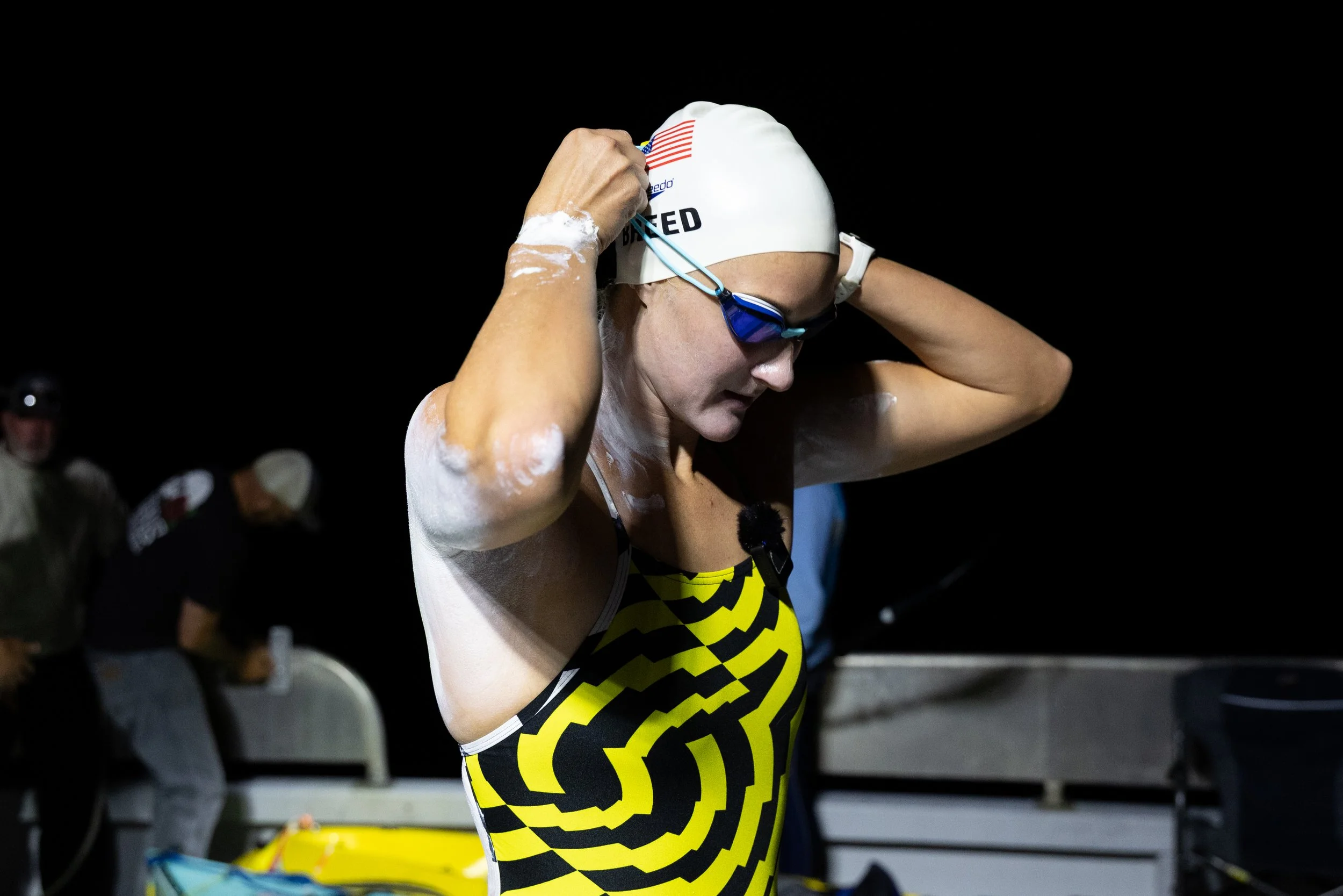 Female swimmer in a yellow and black swimsuit adjusting her swim cap and goggles at night before a race, with others in the background.