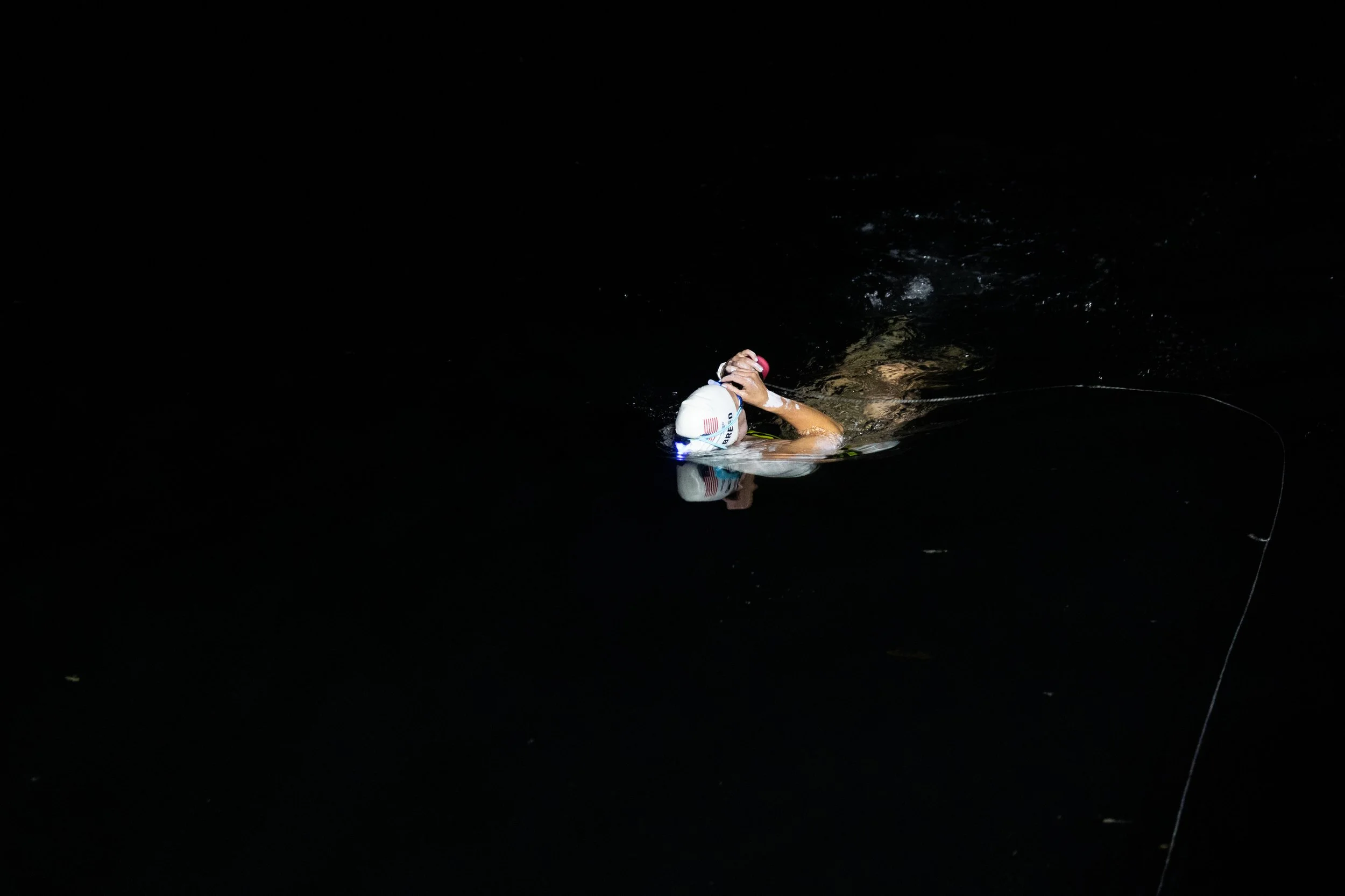 A swimmer in a white cap and goggles swimming in dark water at night.