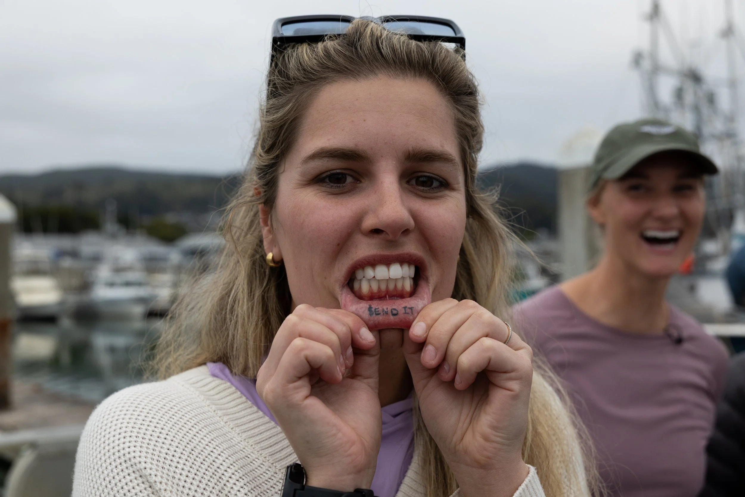 Woman holding a tattooed lip-shaped object with 'SEND IT' written on it, smiling and showing teeth, with another woman smiling in the background at a marina with boats.
