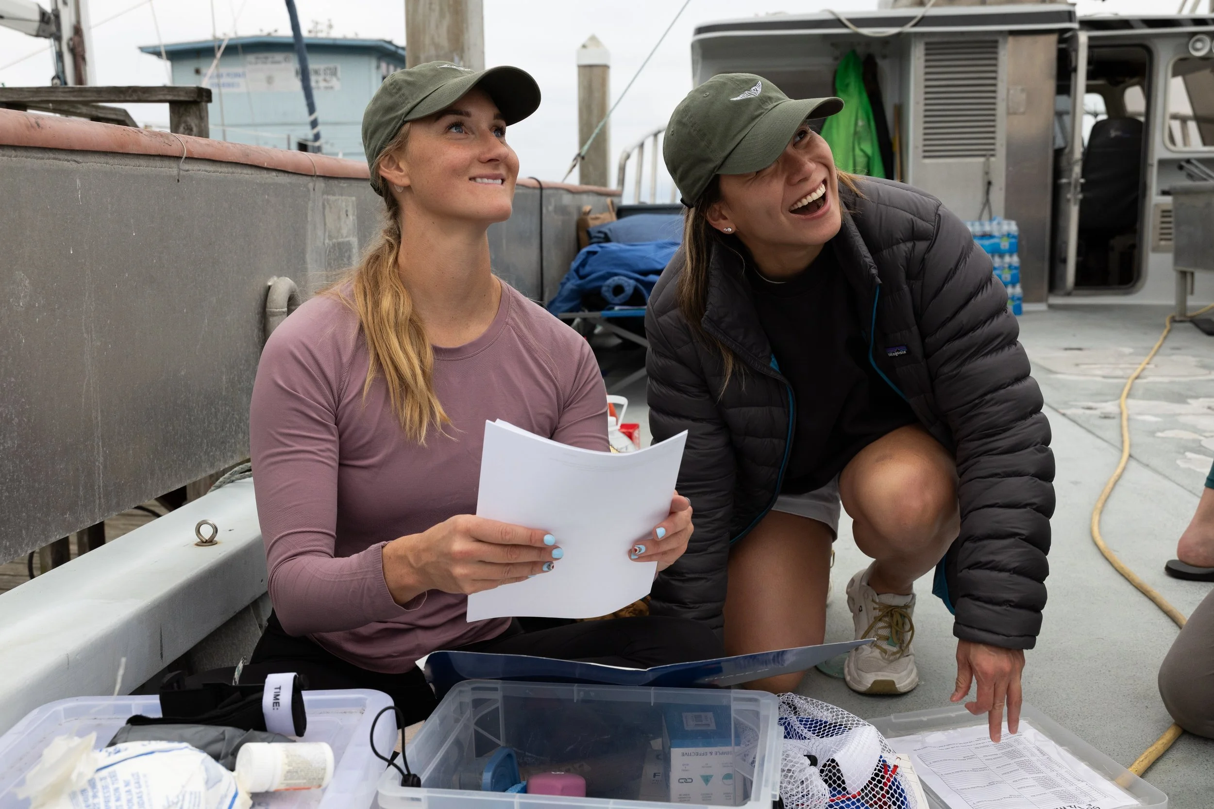 Two women wearing outdoor attire and hats sitting on a boat deck with medical supplies and paperwork, smiling and looking at each other.