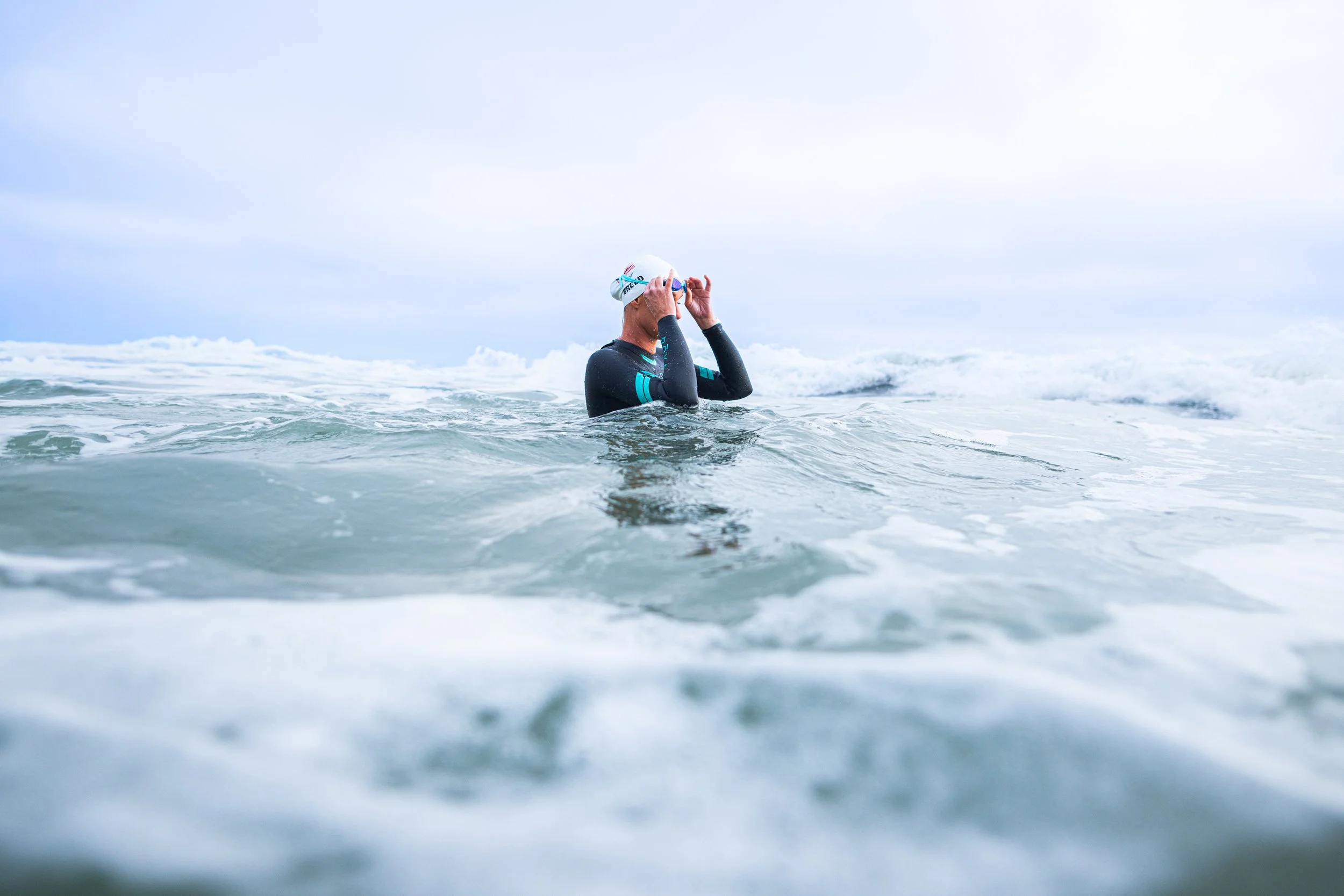A person in a wetsuit is standing in the ocean, adjusting goggles, with waves and cloudy sky in the background.