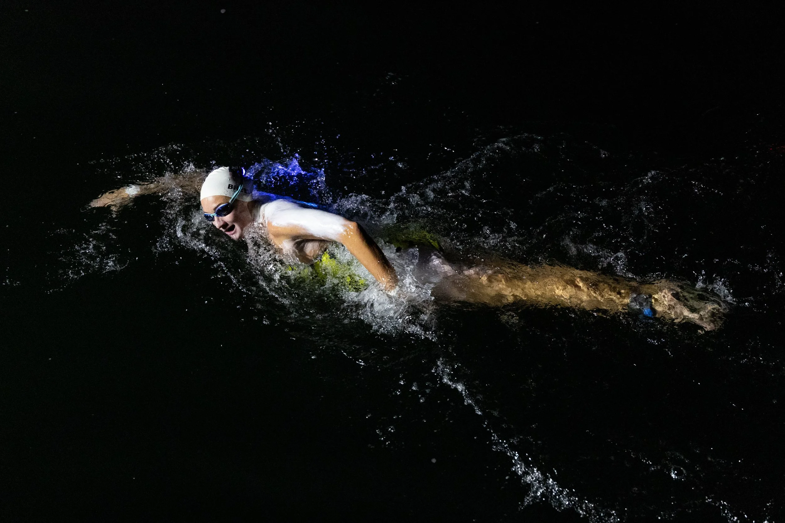 Swimmer in a dark water environment, wearing goggles and a swim cap, actively swimming freestyle.