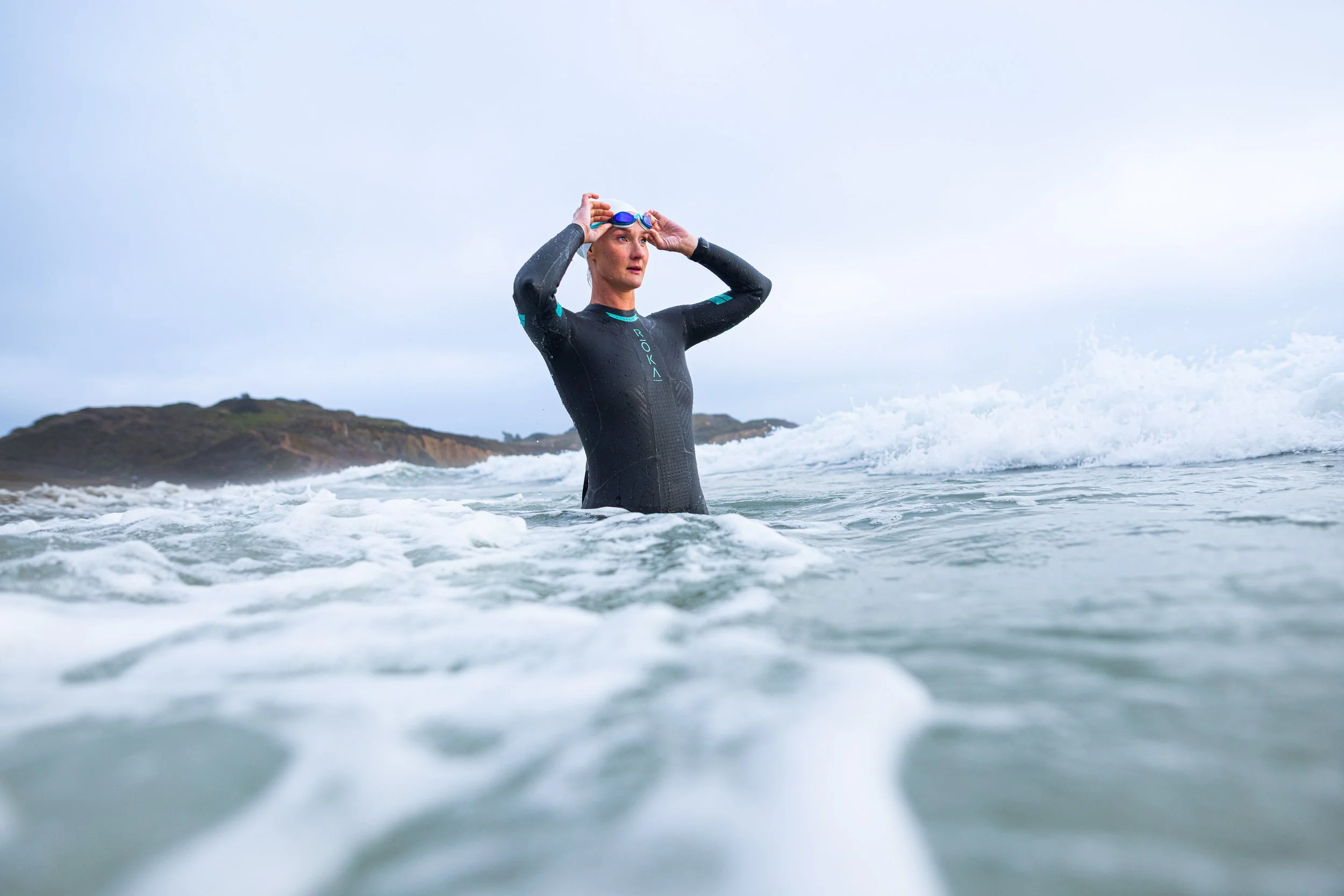 Woman in black wetsuit standing in ocean, adjusting goggles, with island and cloudy sky in background.