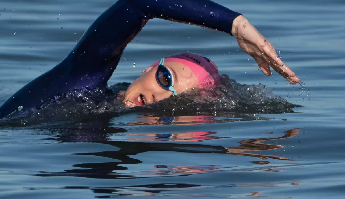 Swimmer in a black wetsuit swimming in open water with pink swim cap and goggles, performing a freestyle stroke.