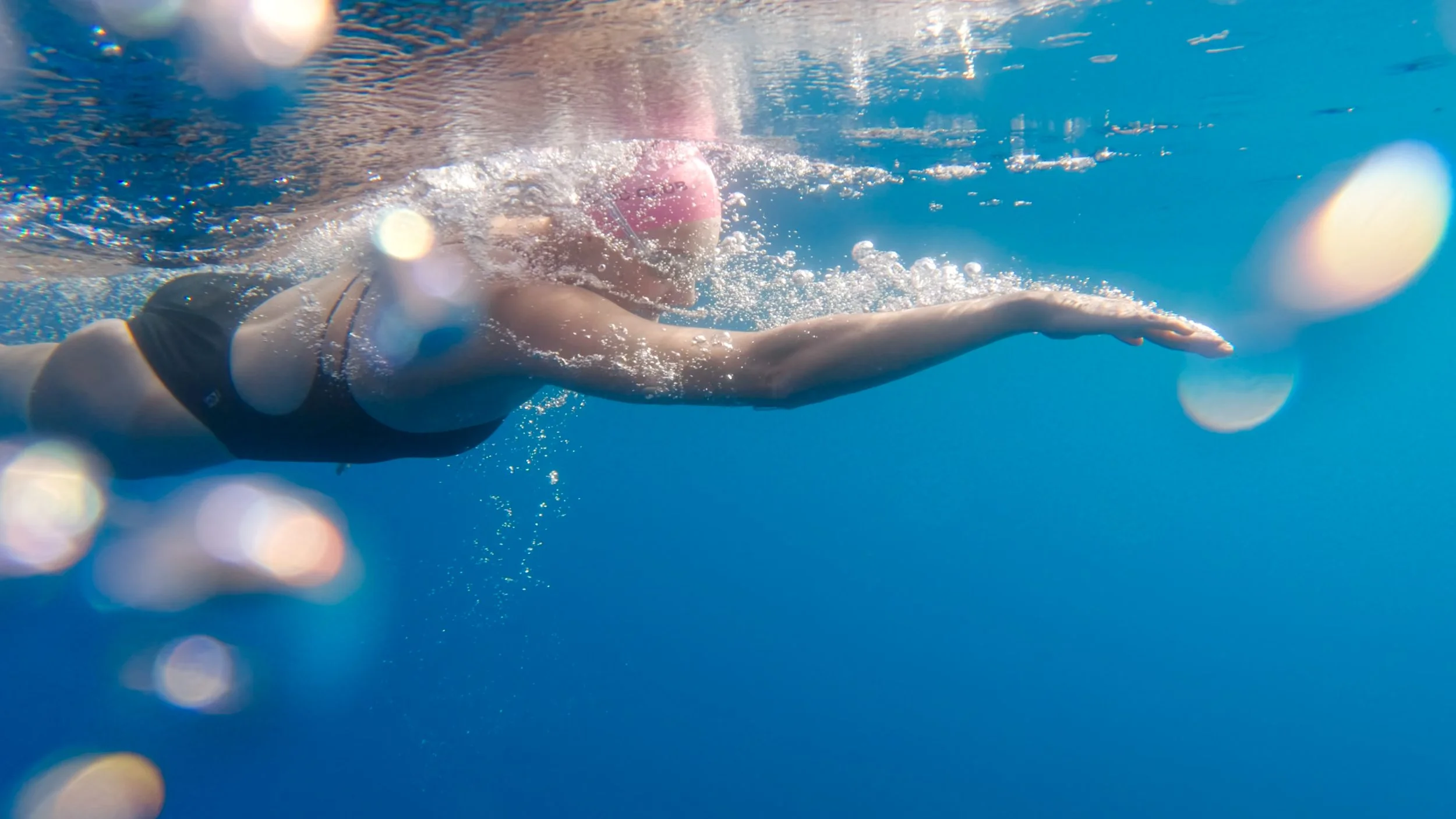 A person swimming underwater in a pool, wearing a pink swim cap and black swimsuit, with an outstretched arm.