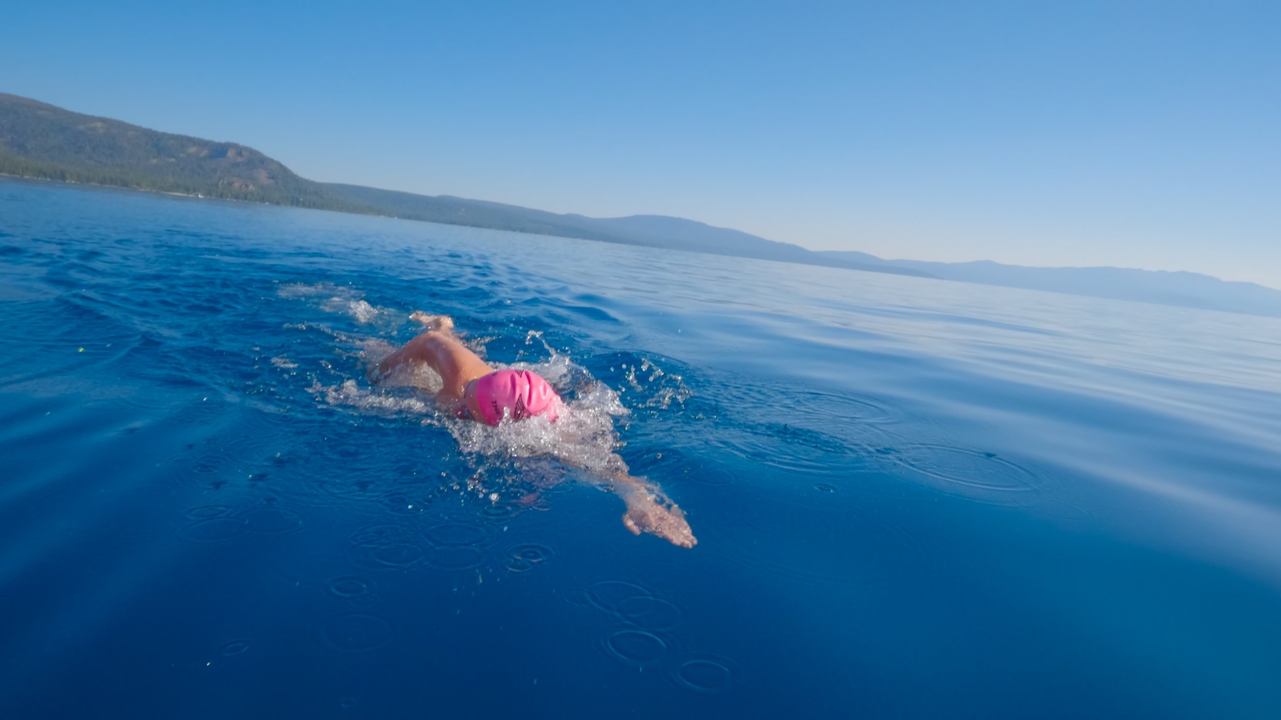 Person swimming in a large body of blue water near a mountainous shoreline on a clear day.