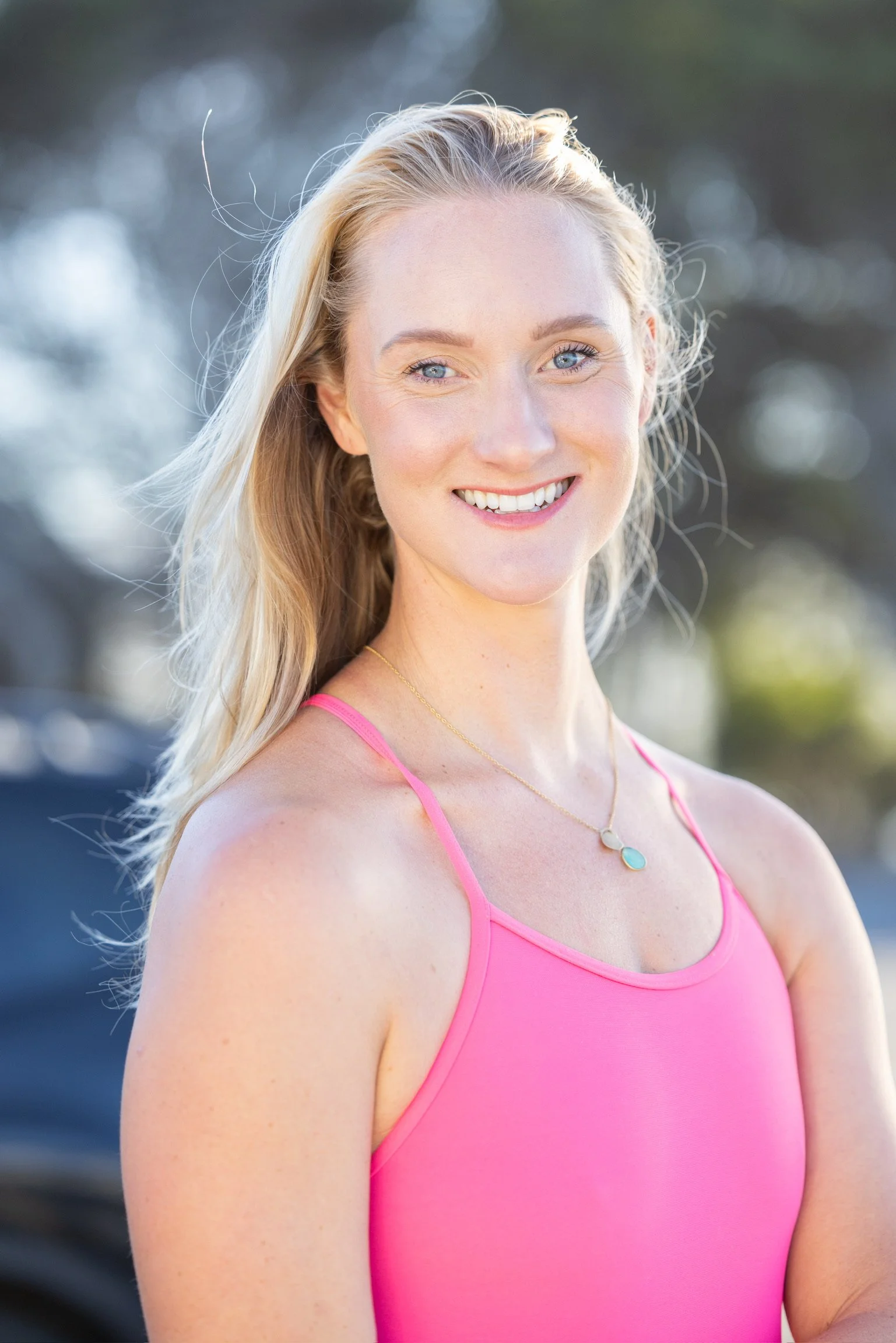 A woman with blonde hair, blue eyes, and fair skin smiling outdoors in natural light, wearing a pink tank top and a delicate pendant necklace.
