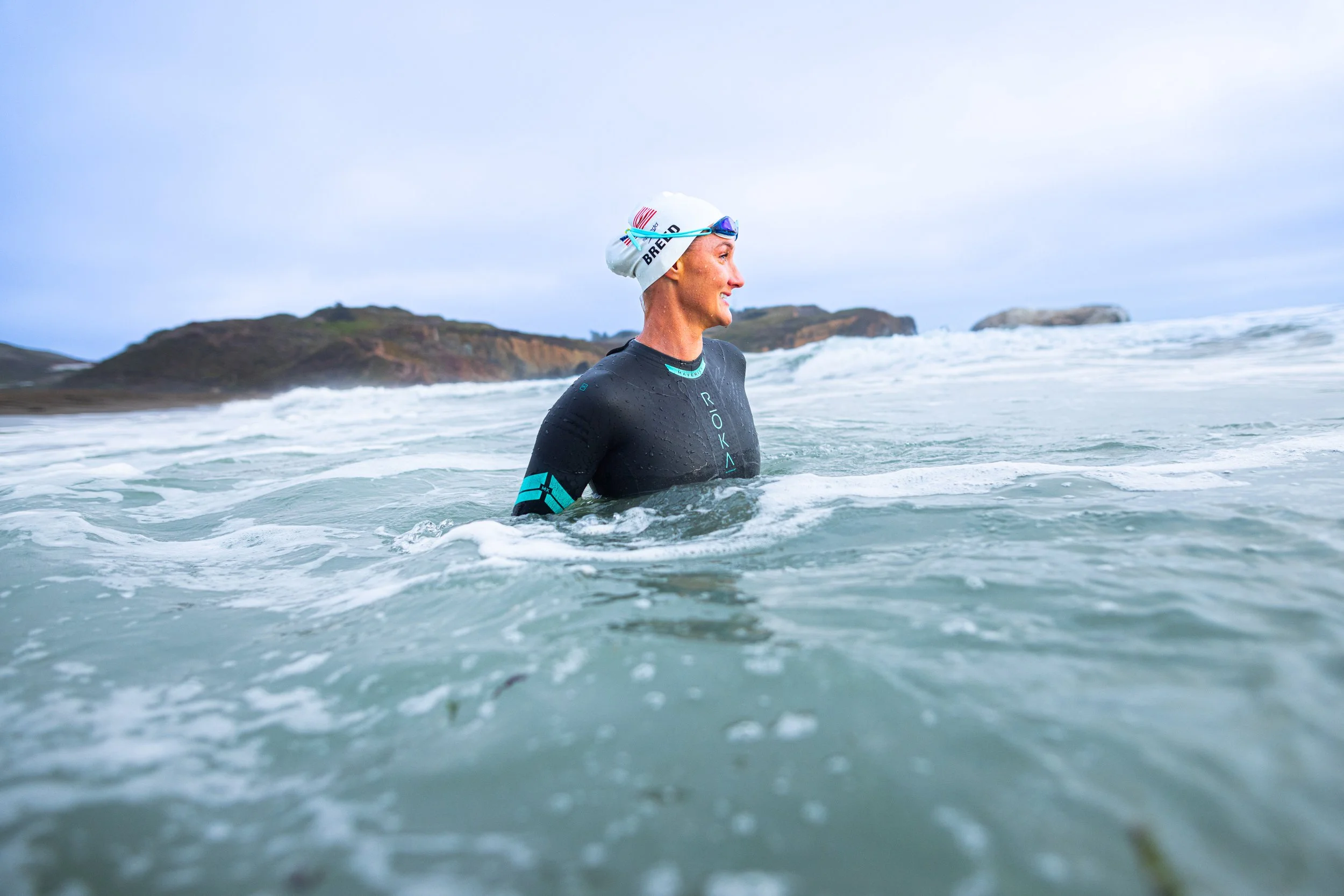 A woman in a black wetsuit with blue accents and a white swim cap standing in the ocean near rocky coastline, smiling and enjoying the water.