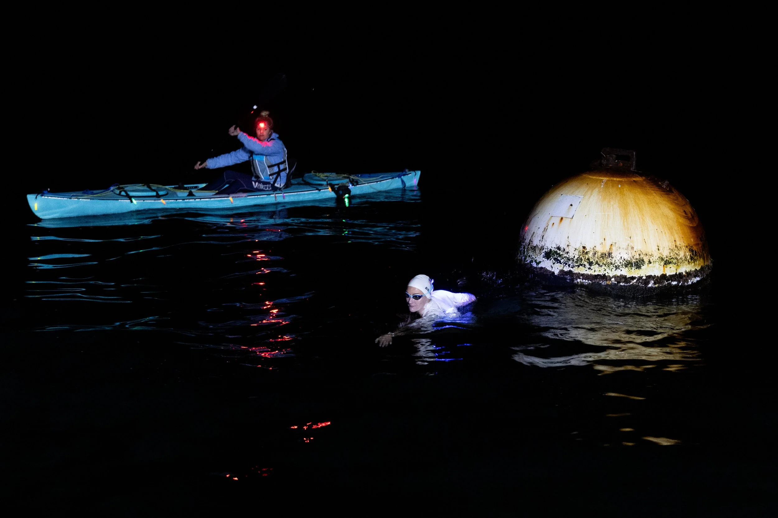 A person in a kayak and another swimmer in dark water at night, near a large, rusted buoy.