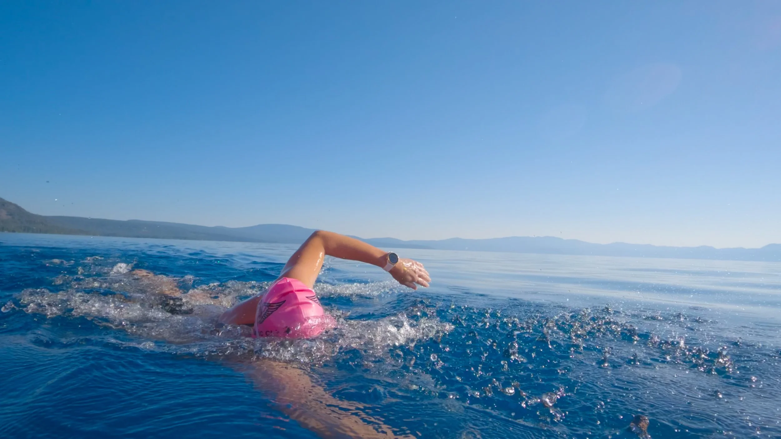 Swimmer in a pink cap and athletic watch swimming in a large body of water with distant mountains under a clear blue sky.
