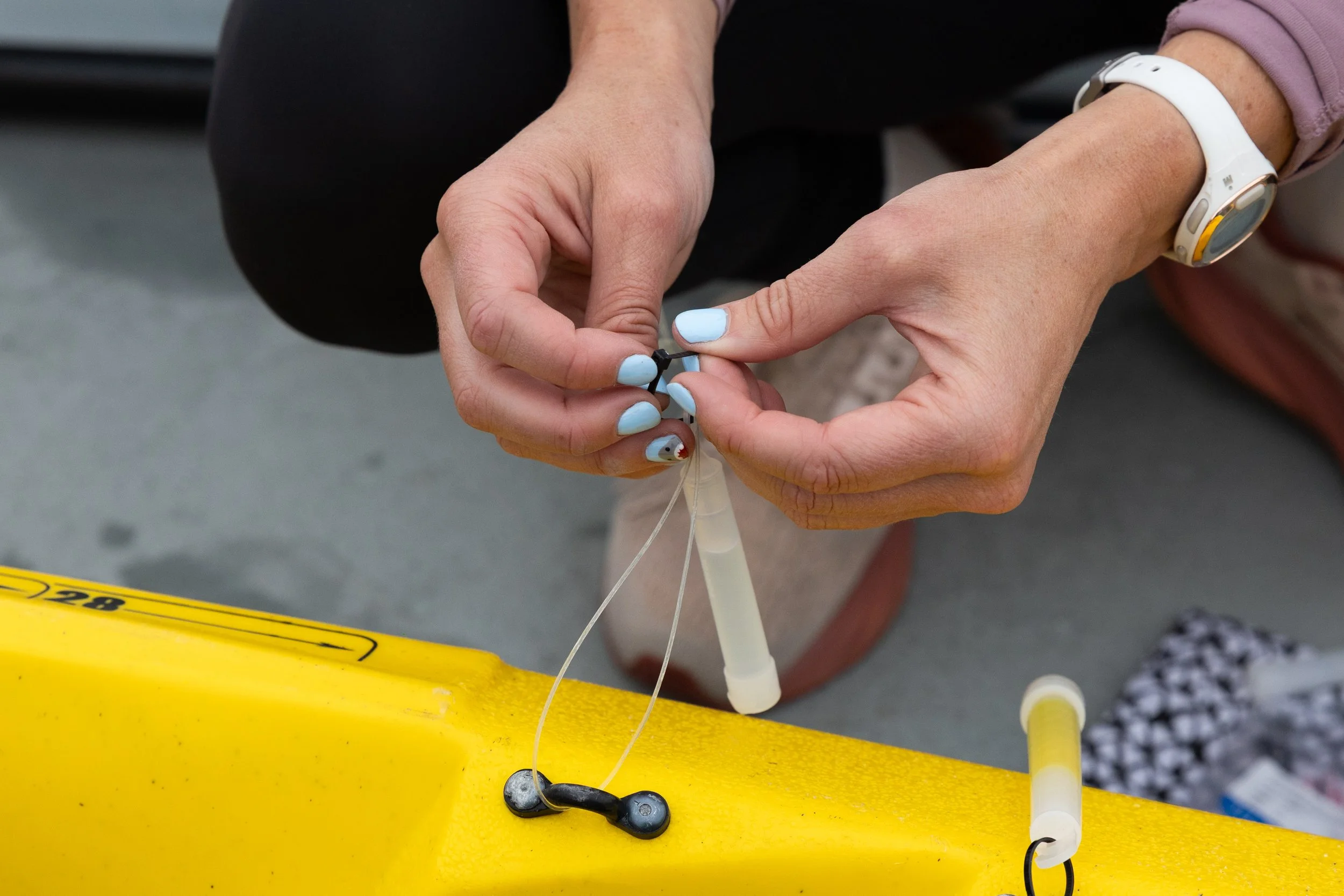 Hands of a person with blue painted nails assembling a piece of medical or scientific equipment with tubing and a syringe, on a yellow surface.