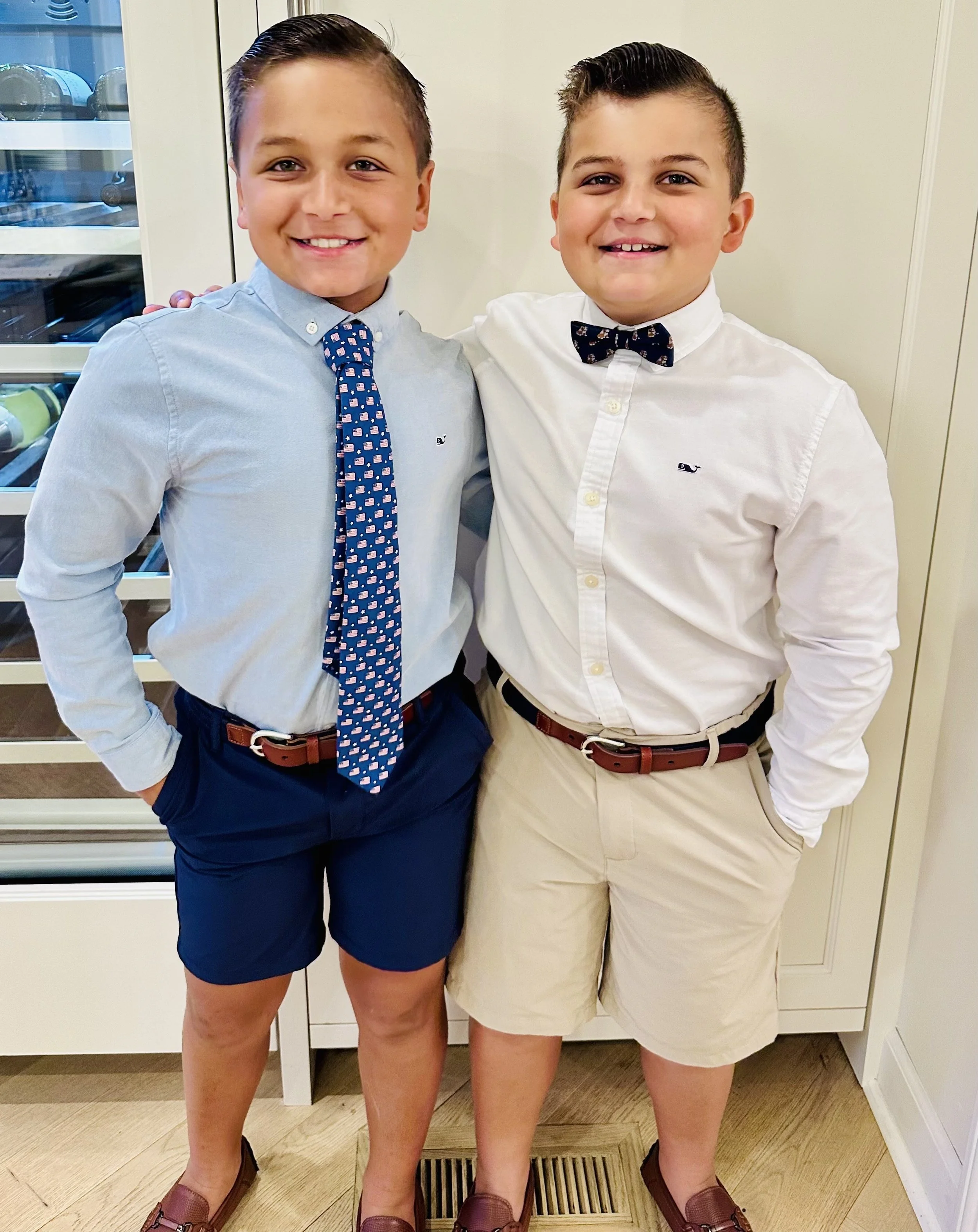 Two young boys standing side by side, smiling, dressed in semi-formal attire. The boy on the left wears a light blue shirt with a patriotic patterned tie, navy shorts, and brown loafers. The boy on the right wears a white shirt with a navy bow tie, beige shorts, and brown loafers. They have their arms around each other's shoulders.