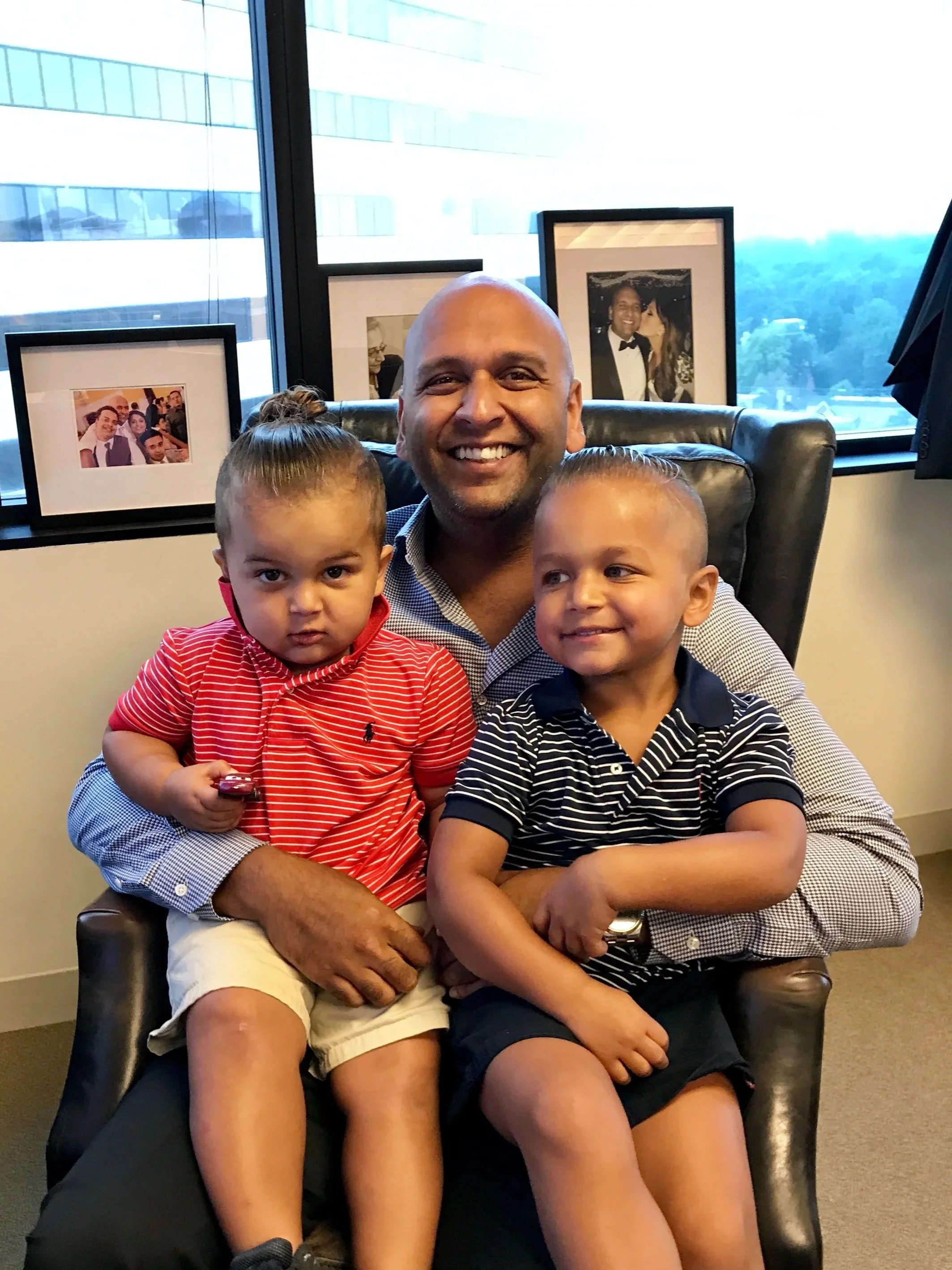 A man smiling and sitting in an office chair with two children on his lap. Behind them are framed photos on a windowsill and a large window showing a cityscape.