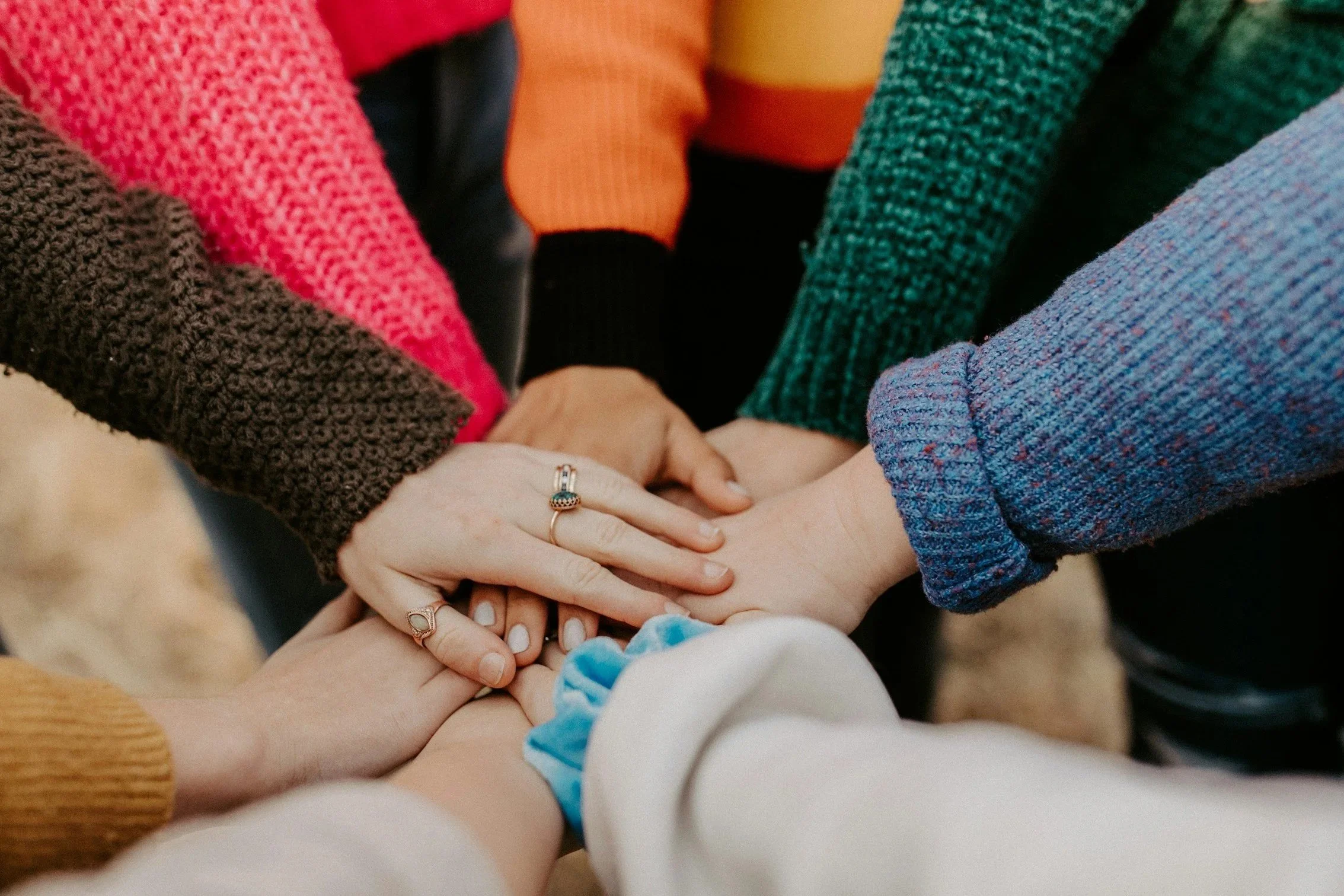 A group of people are stacking their hands together in a show of unity, with varied skin tones and colorful knit sweaters.