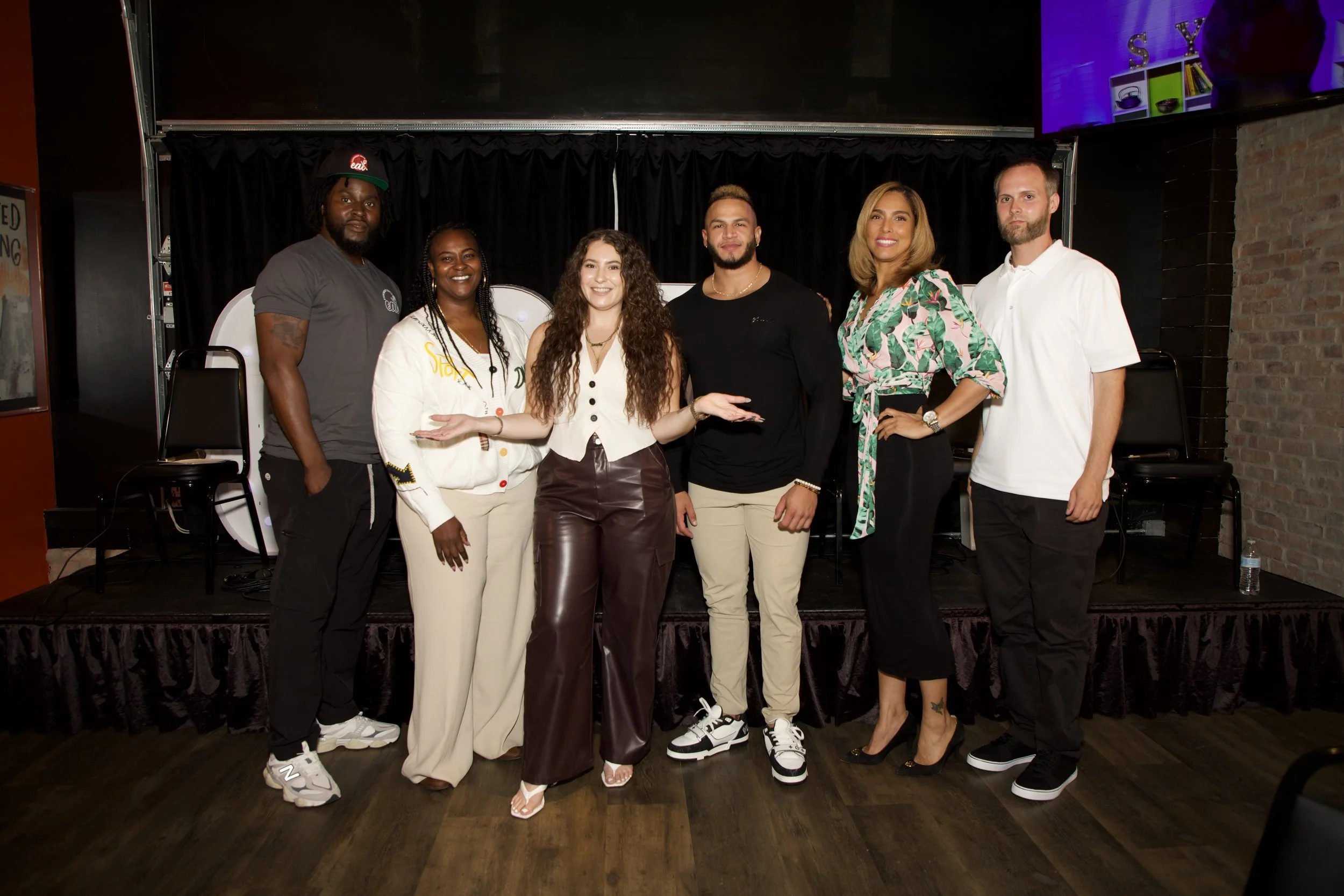 A group of six diverse people standing on a stage with a black curtain background, smiling and posing for the photo. They are dressed casually, with some wearing sneakers and others wearing stylish footwear, and are standing close together for the picture.