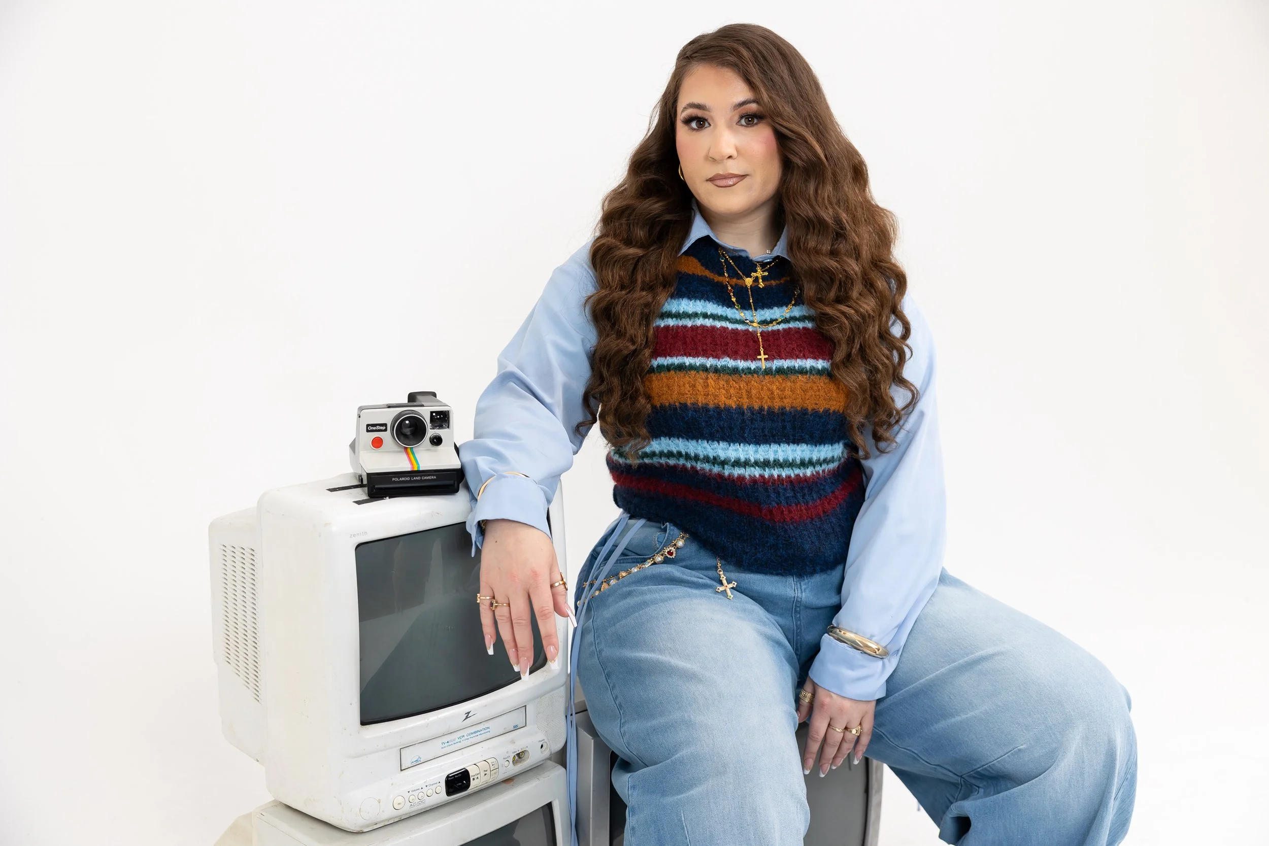 A woman with long, curly brown hair, wearing a multicolored striped sweater over a blue shirt, jewelry including a gold cross necklace, and blue jeans, is sitting on a vintage CRT monitor with a Polaroid camera placed on top, against a plain white background.