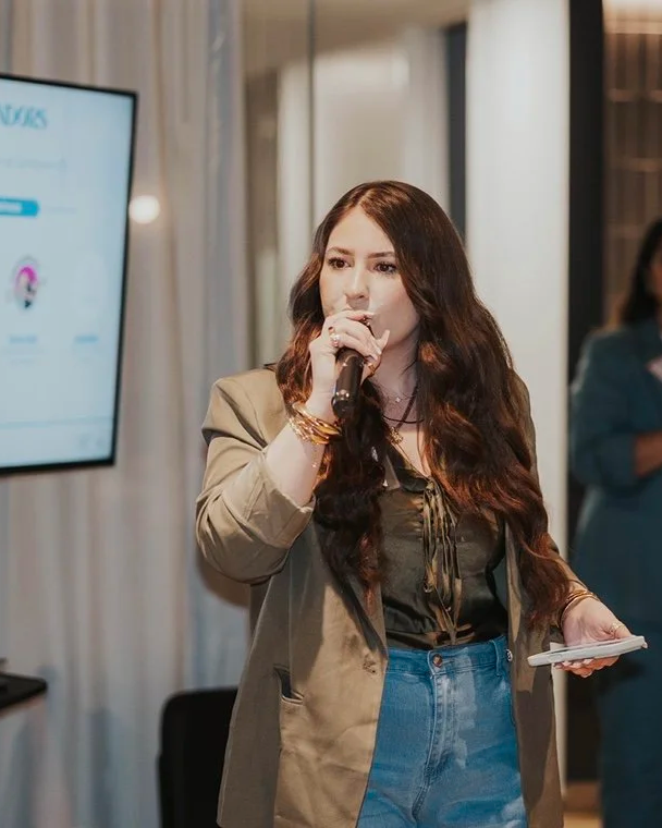 A woman with long wavy brown hair holding a microphone in her right hand, wearing a beige blazer, dark top, and blue jeans, speaking in front of a presentation screen in an indoor setting.