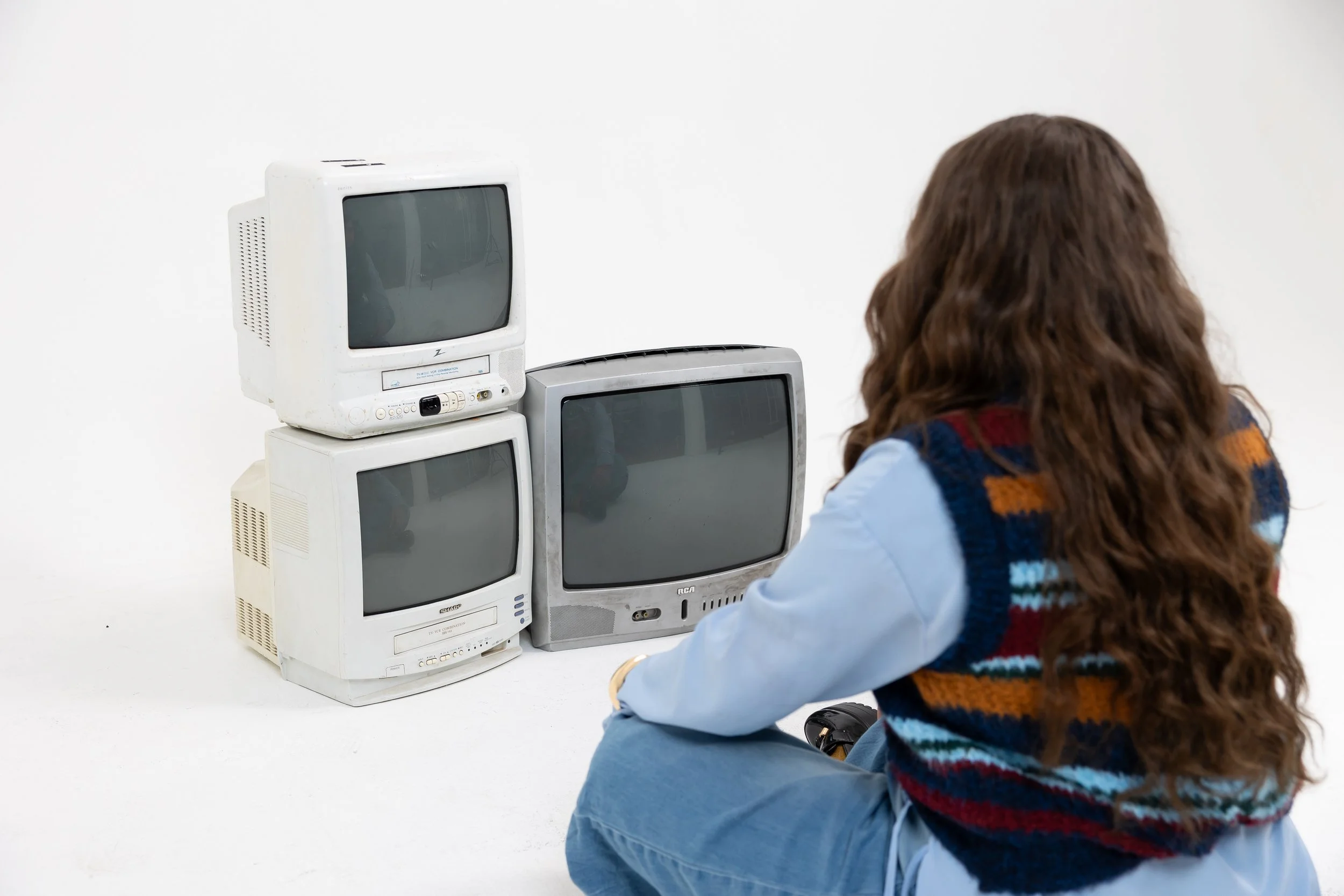 A person with long, curly brown hair wearing a blue shirt and a colorful striped vest sitting on the floor and facing a stack of four old television sets against a plain white wall.
