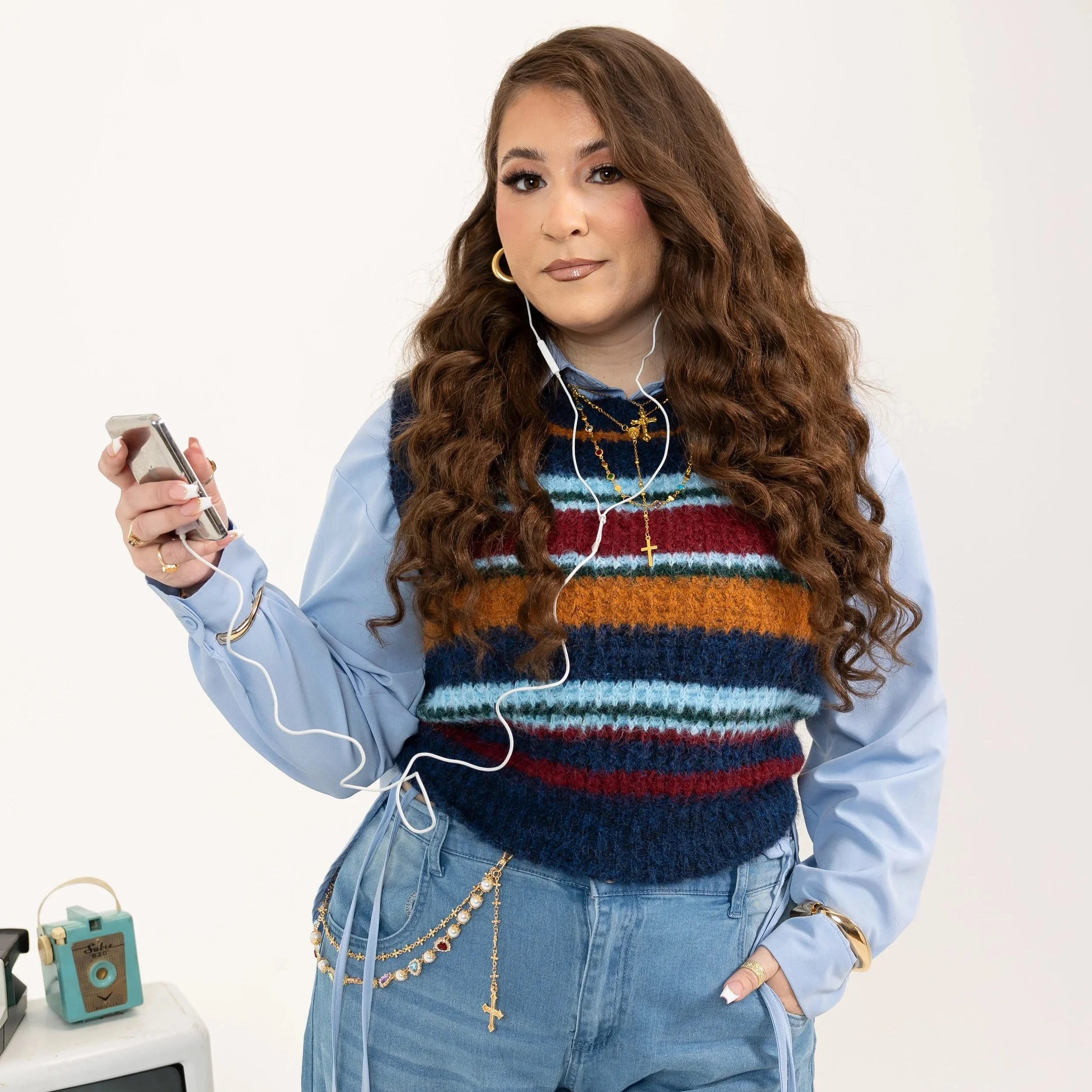 A woman with long curly brown hair, wearing gold jewelry including hoop earrings, layered necklaces with a cross pendant, and rings, is looking at a smartphone while listening to earphones. She is dressed in a light blue jacket over a colorful stripe