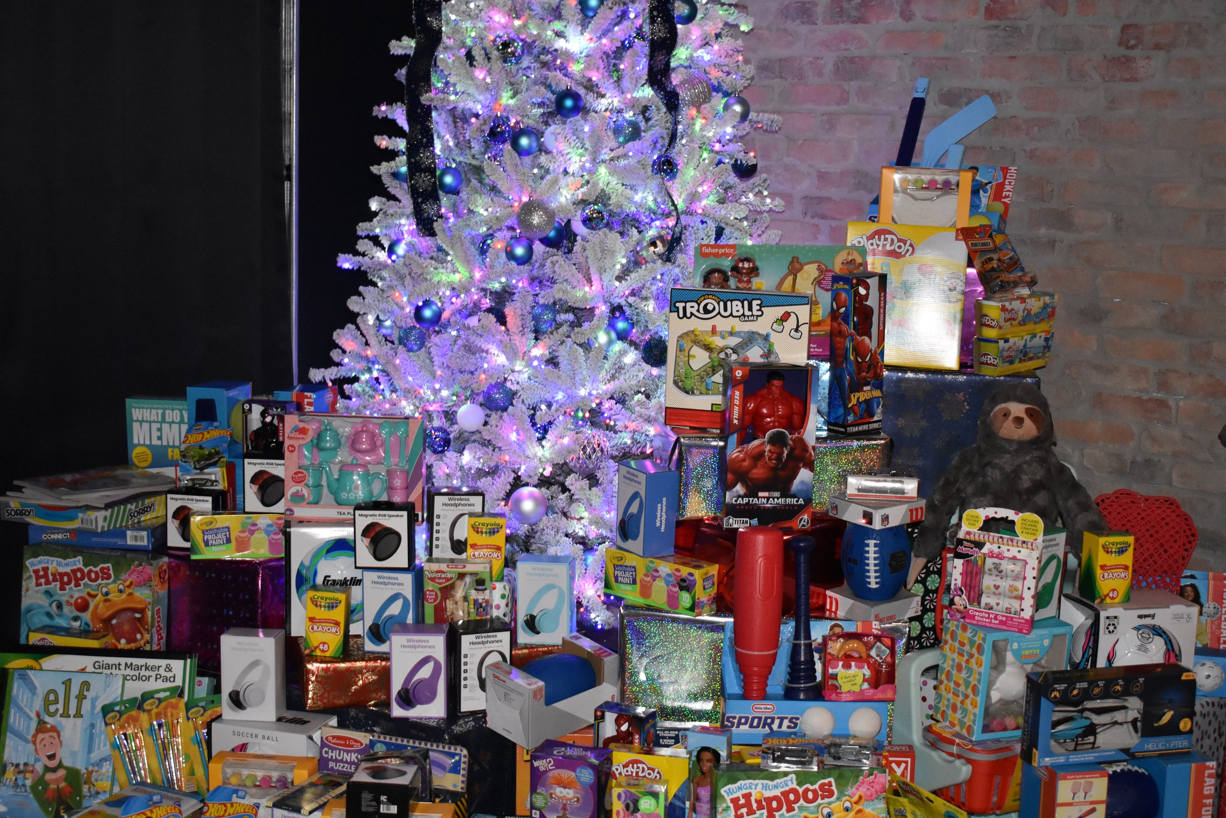 A white Christmas tree decorated with colorful lights and blue ornaments, surrounded by various toys and gifts, against a brick wall background.