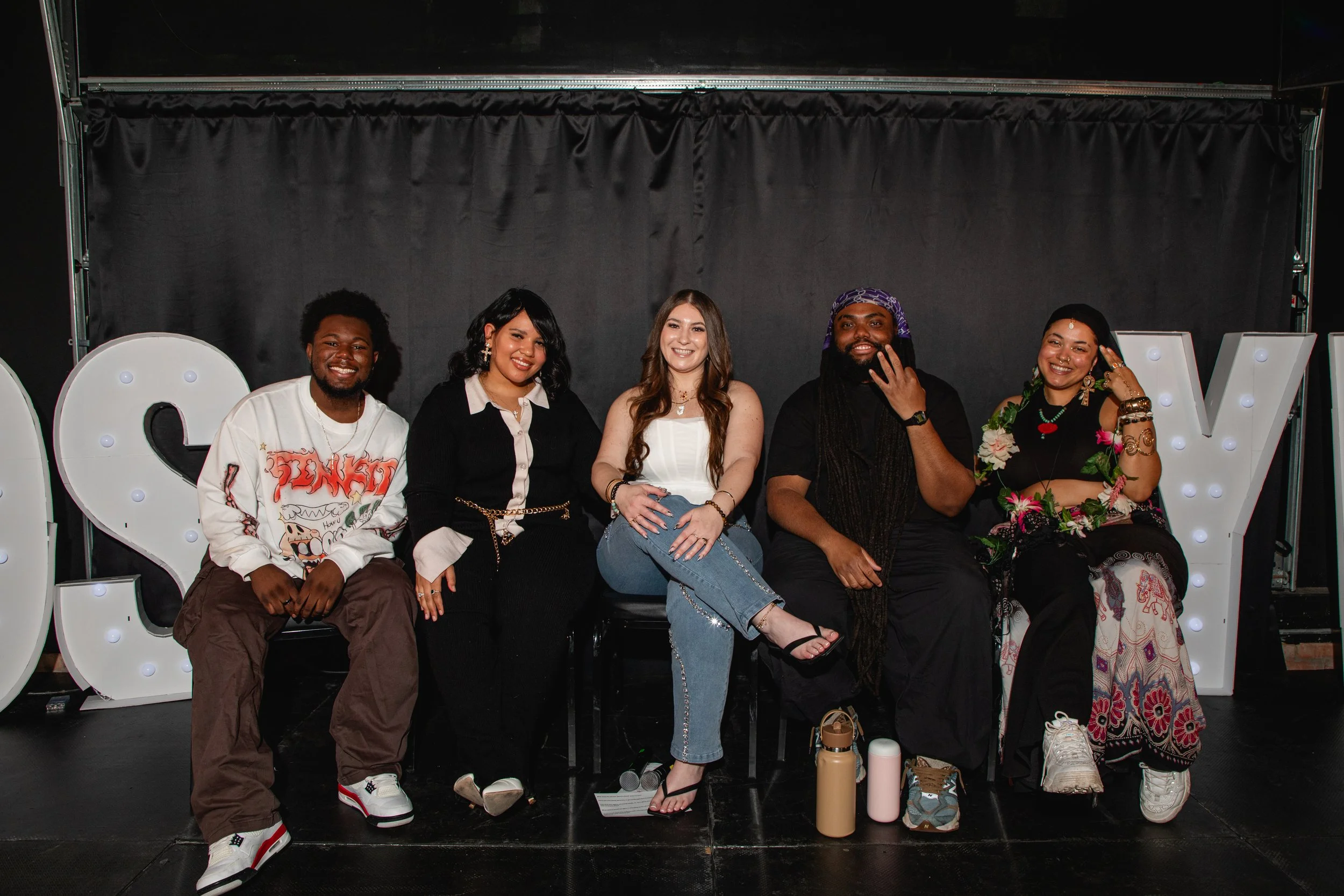 A group of six diverse people sitting on a stage, smiling for a photo during an event, with decorative letters spelling 'LOVE' and personal items on the floor.
