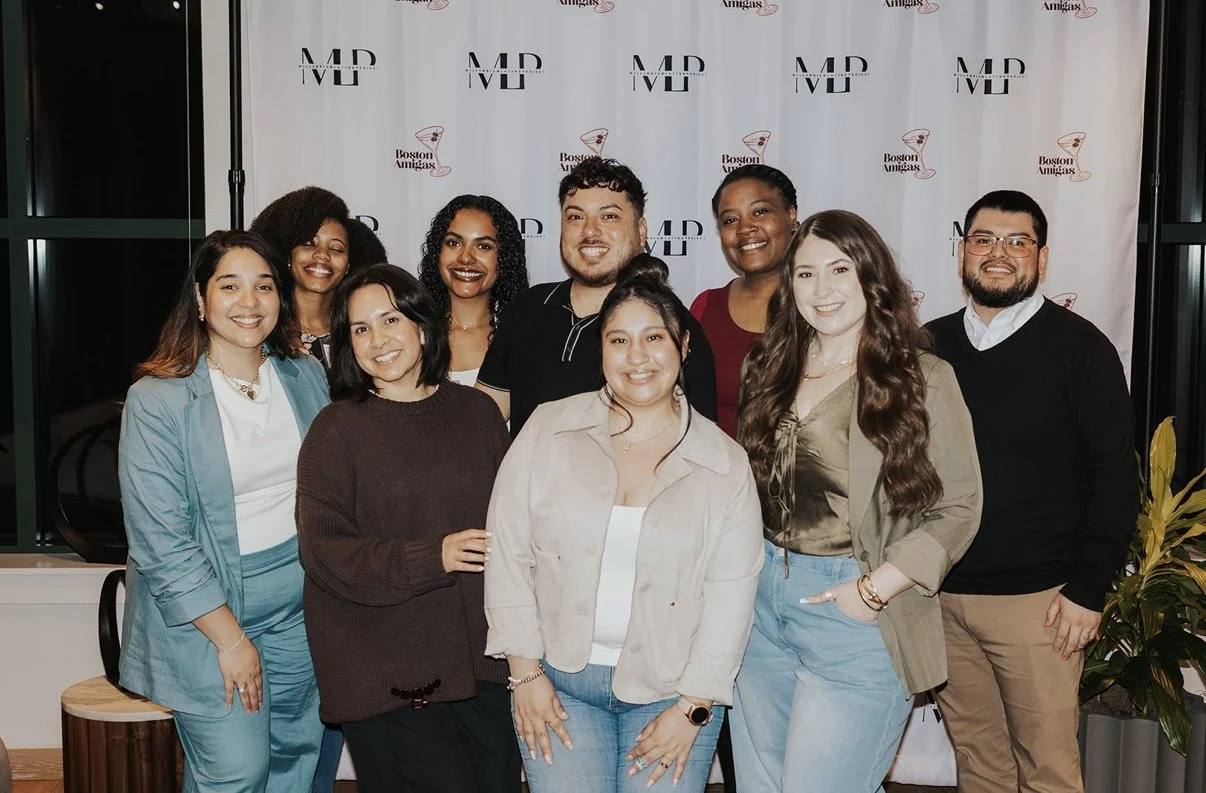 Group of nine diverse people smiling for photo in front of a backdrop with 'MD' and 'Boston Amigas' logos.