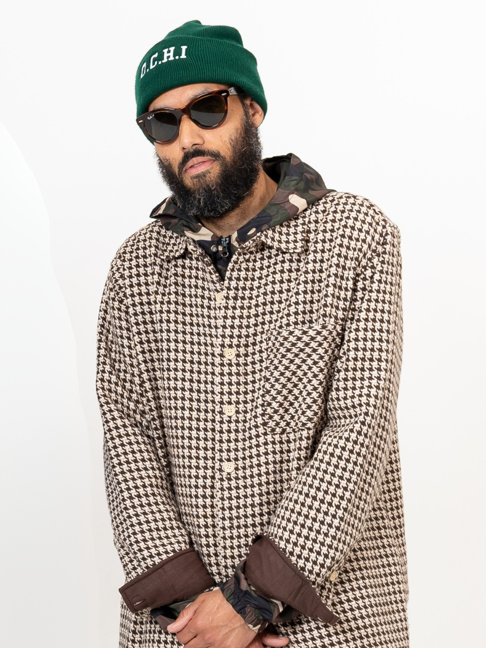 A man with a beard wearing a green beanie, sunglasses, a houndstooth patterned coat, and a camouflage hooded jacket underneath, standing against a plain white background.