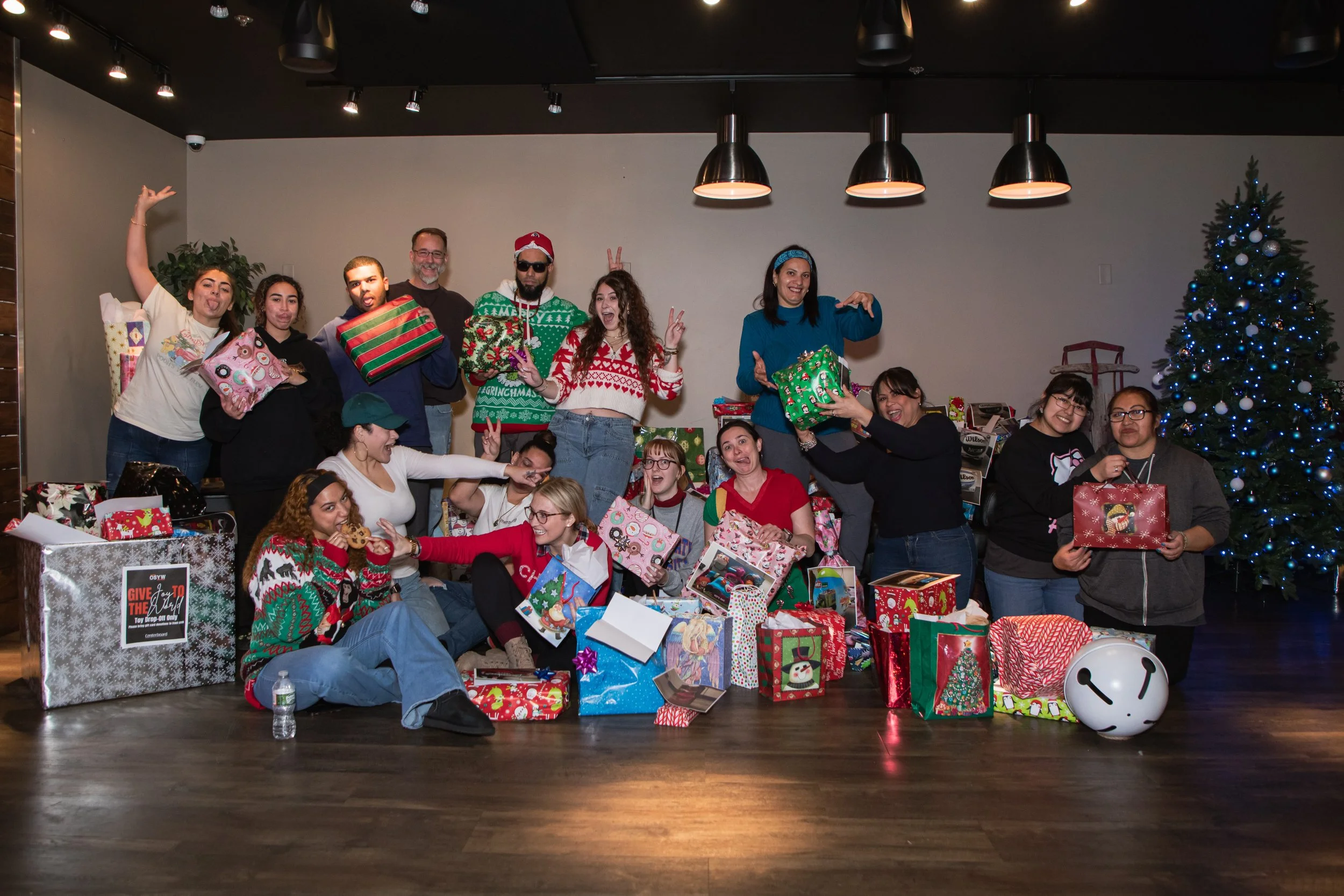 A group of people celebrating a Christmas party with many gifts, a decorated Christmas tree, and festive sweaters, some making funny faces and gestures.