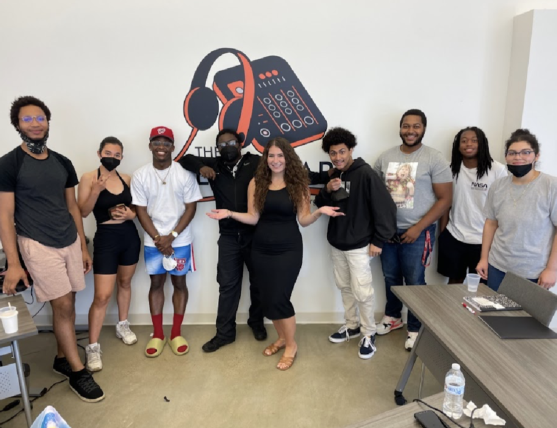 Group of nine people standing indoors in front of a wall with a logo of headphones and a DJ mixer. They are smiling, some wearing masks, and appear to be in a casual or creative setting.