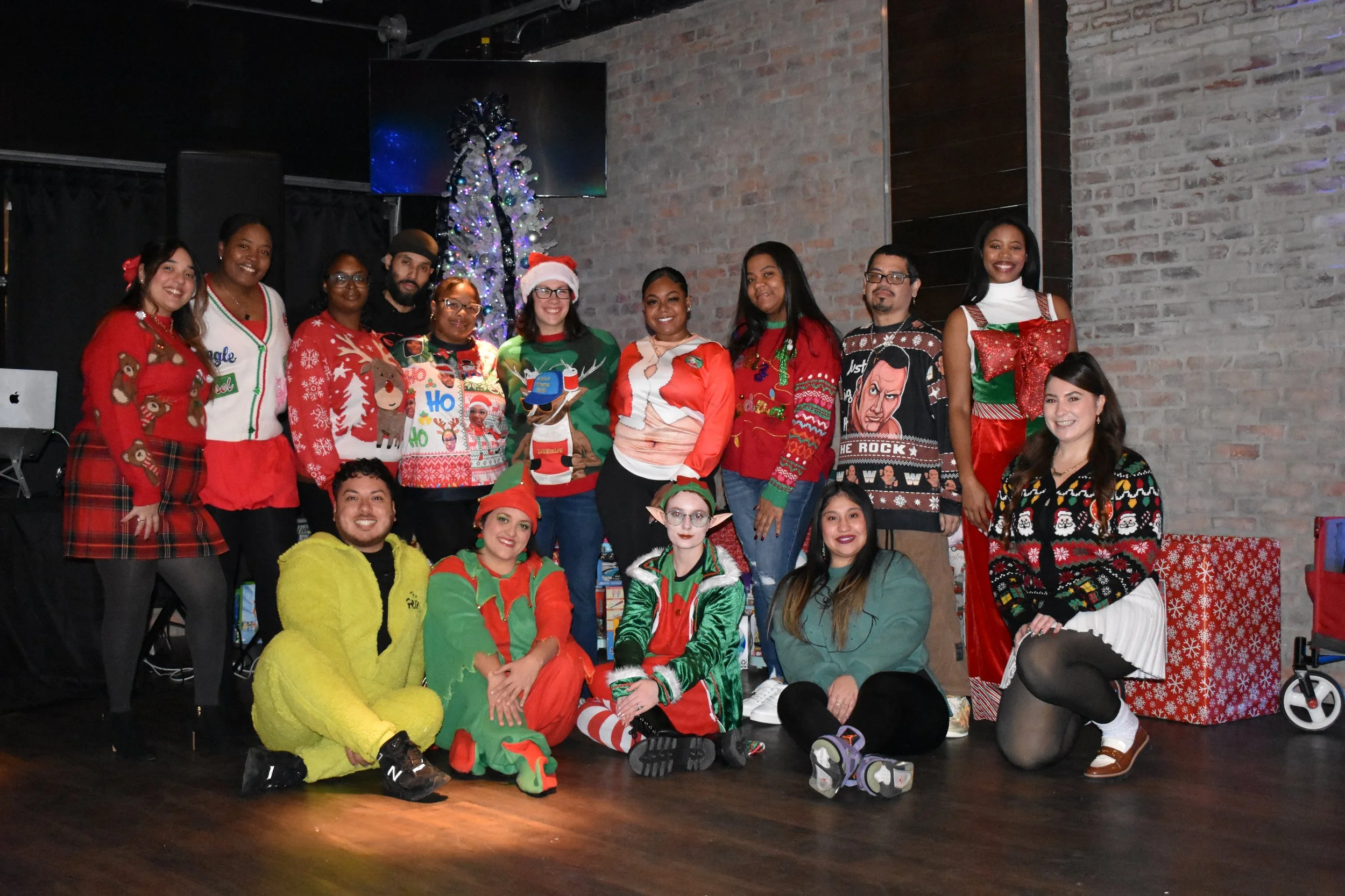 Group of people dressed in Christmas sweaters and costumes, posing in front of a decorated Christmas tree and a brick wall.