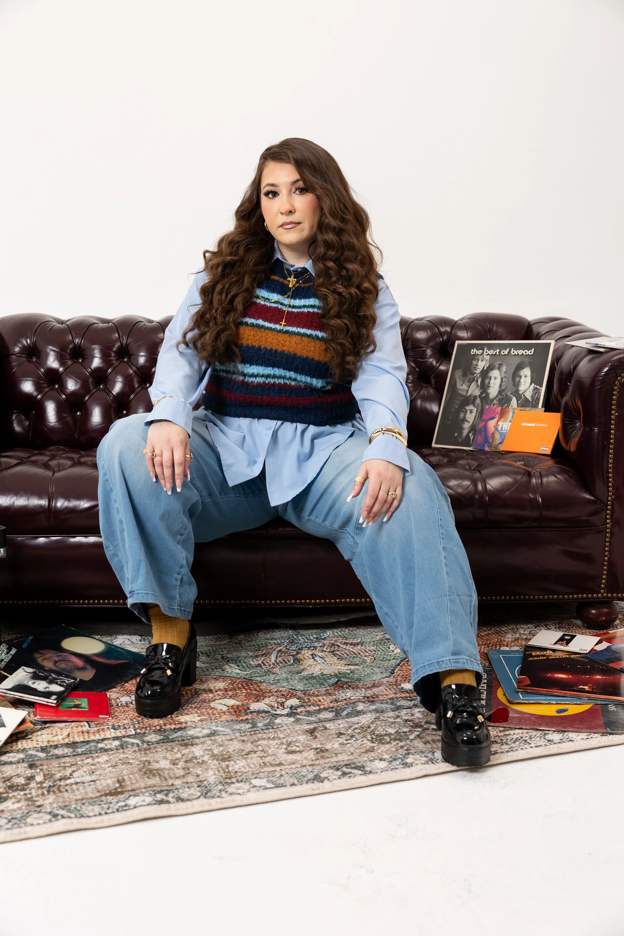 Young woman sitting on a vintage leather sofa surrounded by vinyl records and music albums, wearing a colorful striped sweater and loose jeans.