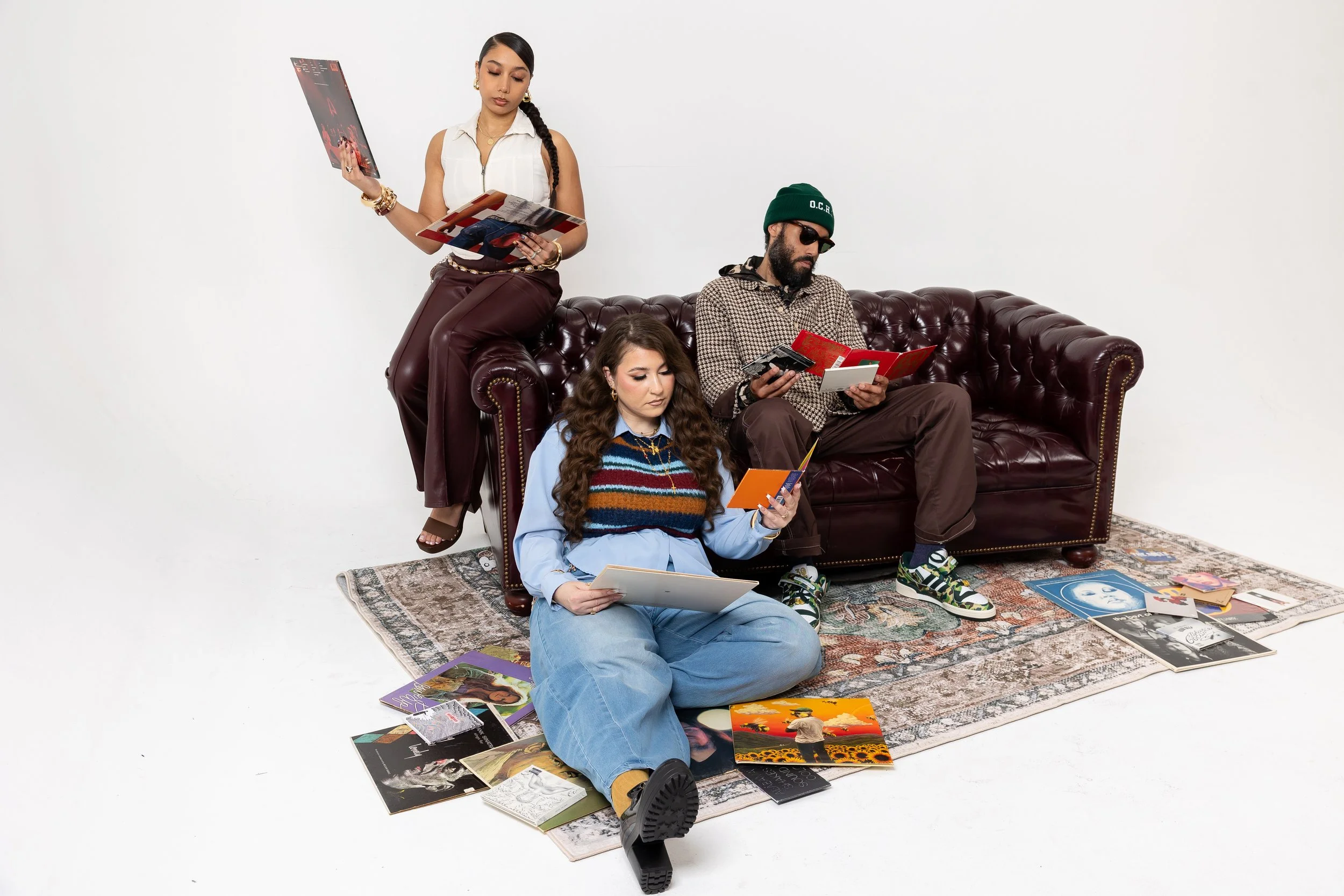 A group of three young adults, two women and one man, in a minimalistic white room with a decorative rug, surrounded by various magazines and posters, engaging with printed materials.