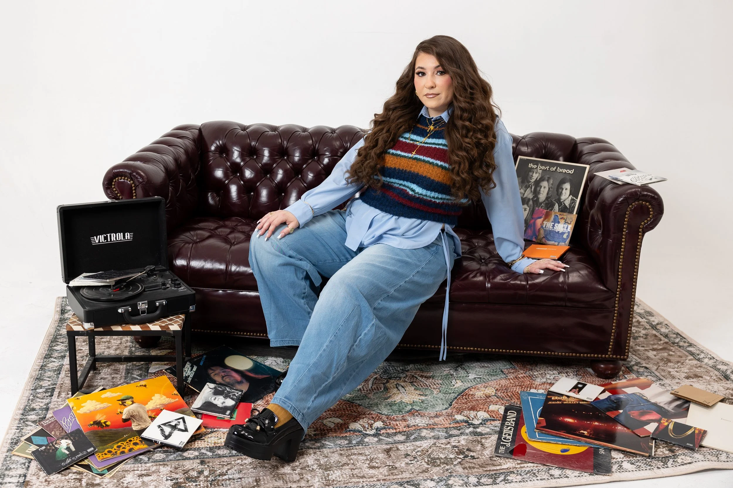 A woman with long curly brown hair, wearing a blue shirt, colorful striped sweater vest, and blue jeans, sitting on a leather couch surrounded by vinyl records and album covers with a turntable nearby.