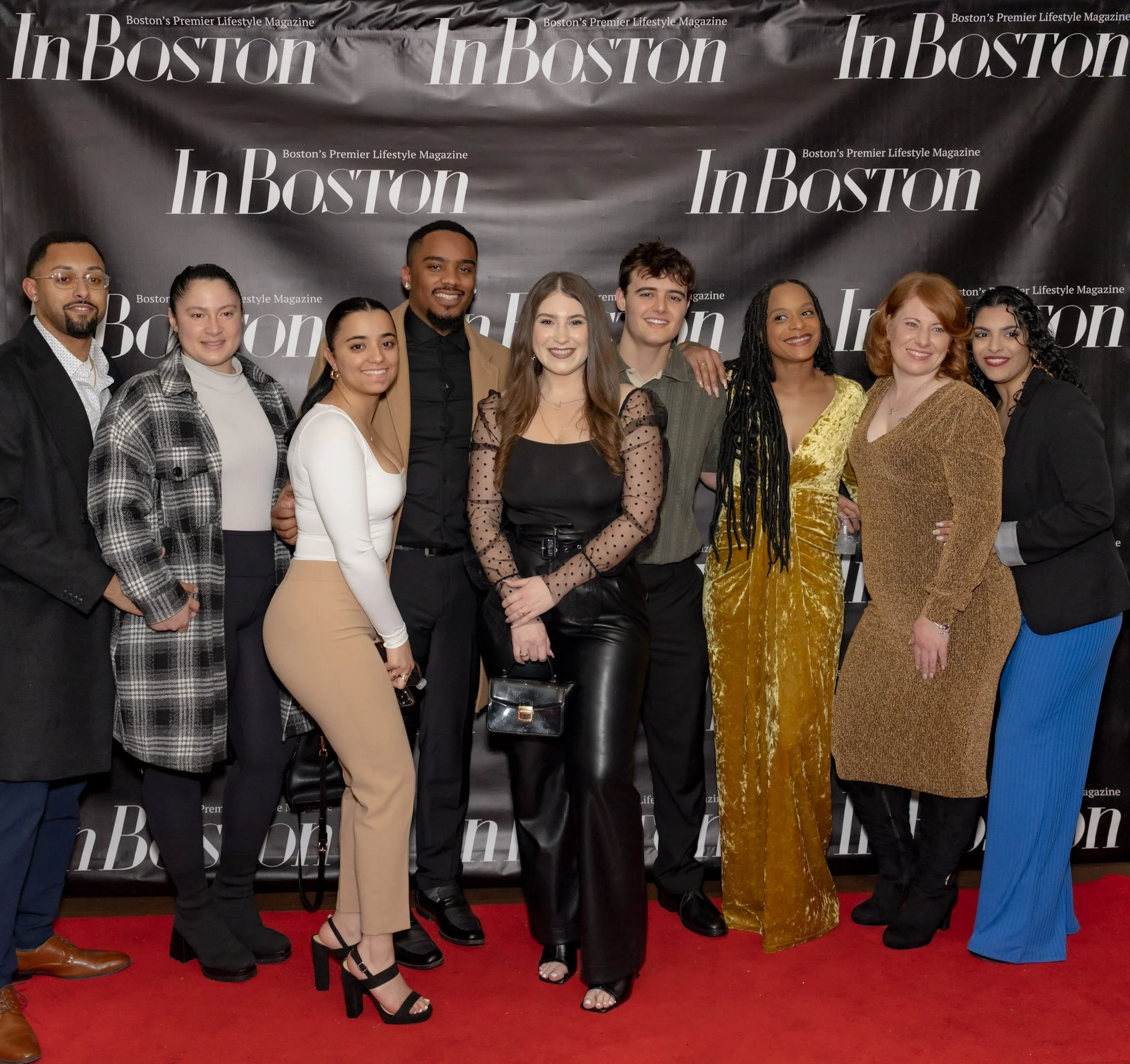 Group of ten diverse young adults standing on a red carpet in front of a black backdrop with 'In Boston' and 'Boston's Premier Lifestyle Magazine' printed repeatedly. They are smiling, dressed in stylish clothing, and appear to be attending an event or celebration.