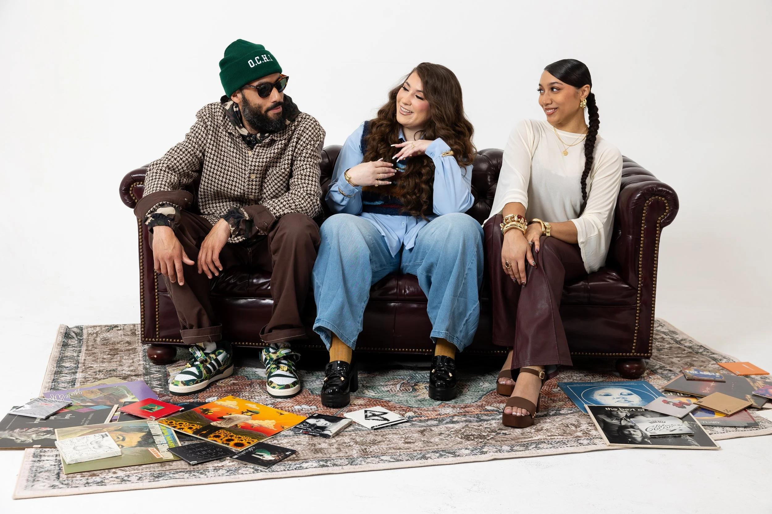 Three women sitting on a leather couch in a white room surrounded by magazines scattered on the floor. They are engaged in conversation, with the woman in the middle smiling and the women on either side looking at her. The woman on the left is wearin