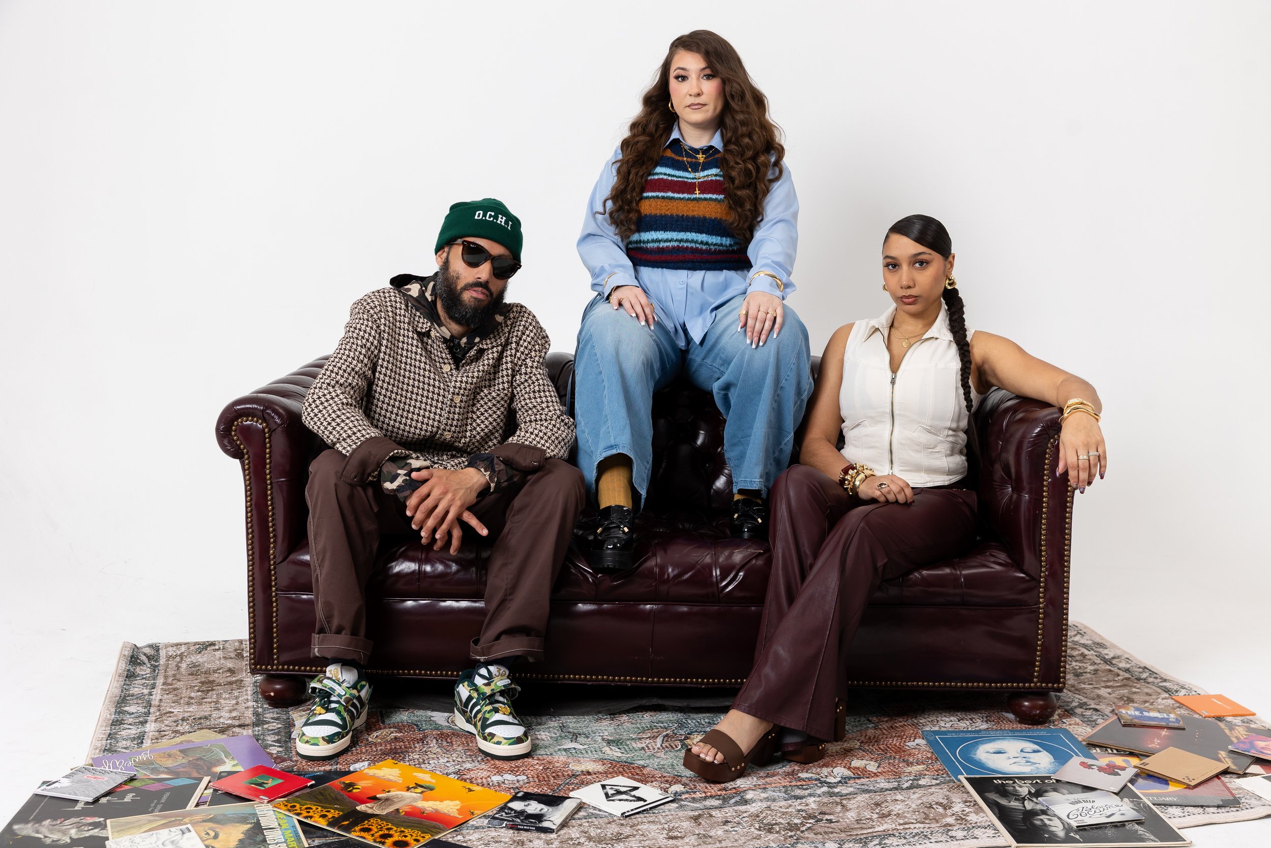 Three people sitting and standing on a vintage leather couch covered with magazines spread on a patterned rug in a plain white room.