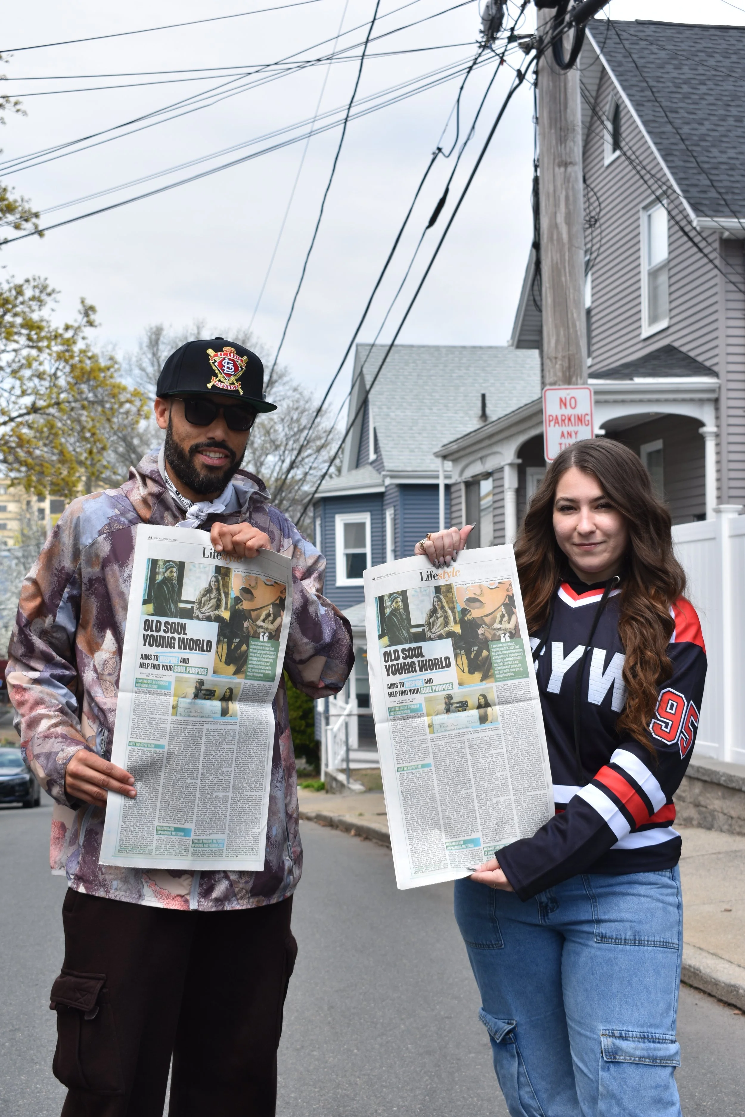 Two people standing on a street holding newspapers with a headline about old soul and young world. The man on the left is wearing sunglasses, a black cap, and a camouflage jacket. The woman on the right has long brown hair, a black and red sports jersey, and blue jeans. There are houses and a utility pole with wires in the background.