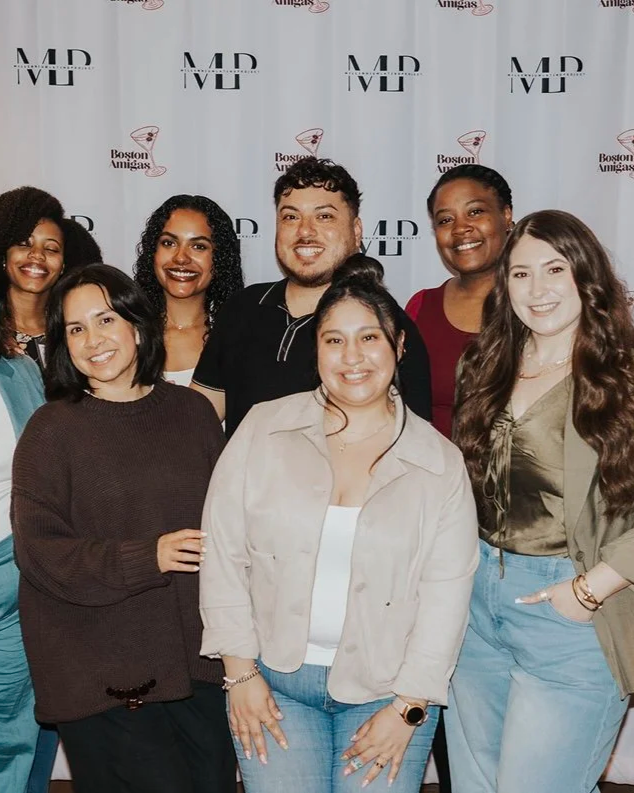 Group of seven diverse women and one man posing together at an event, standing in front of a backdrop with logos that read 'MD Live' and 'Boston Amigas'.