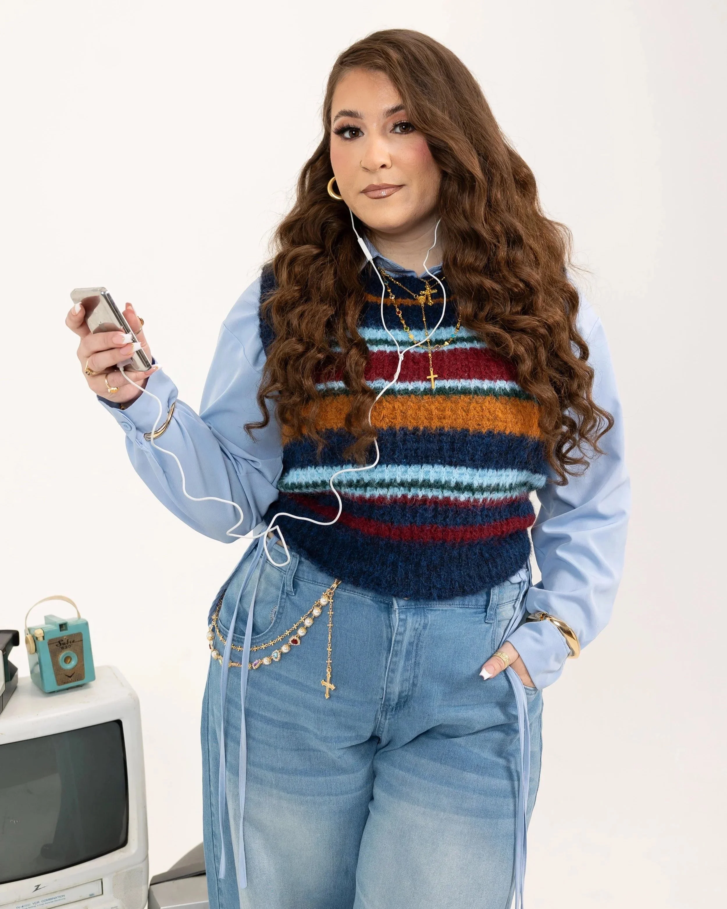 A woman with long, wavy brown hair wearing a blue and multicolored striped sweater layered over a light blue shirt, accessorized with gold jewelry including a cross necklace and hoop earrings. She is holding a smartphone with white earphones plugged in, standing against a plain white background. Part of a small table with a vintage teal camera and a vintage TV are visible in the lower left corner.