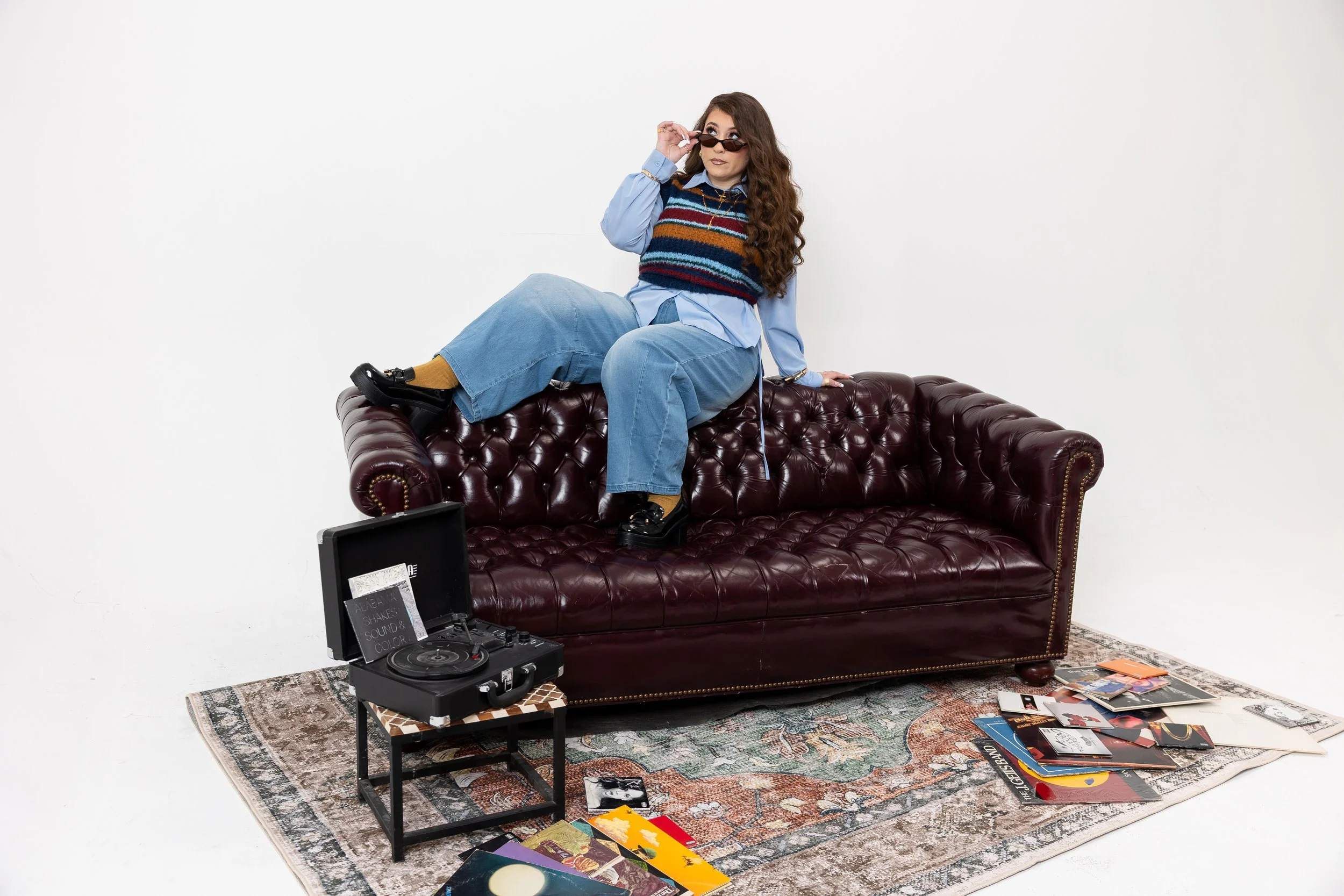 A woman with long curly hair, wearing a striped sweater, blue shirt, and sunglasses, sitting on a vintage leather couch in a room with a plain white wall. There are vinyl records and magazines scattered on the floor, and a record player on a small ta