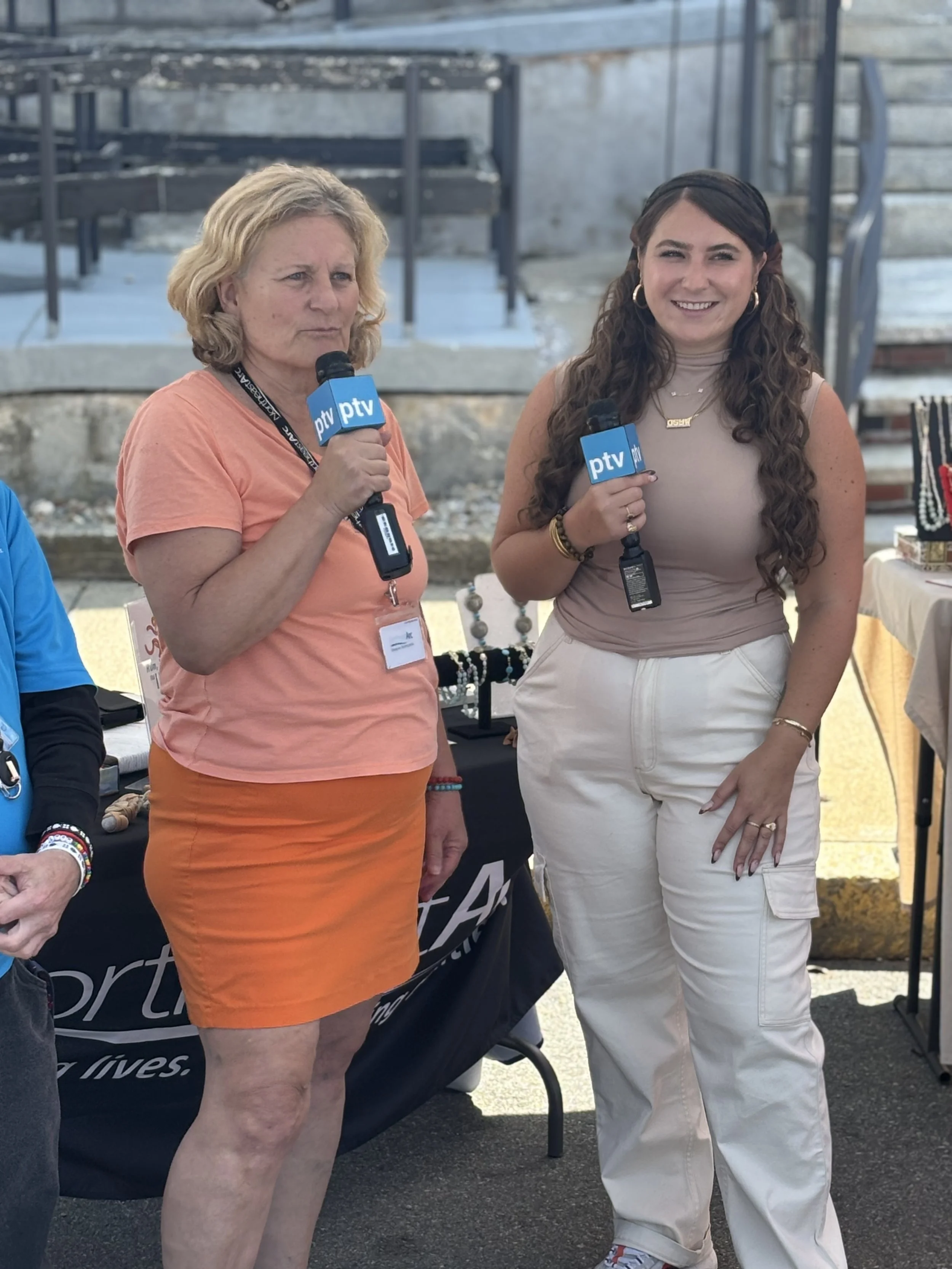 Two women standing outdoors holding microphones labeled 'PTV'. One woman has curly, long brown hair, wearing a beige sleeveless top and white pants. The other woman has short, curly blonde hair, wearing an orange t-shirt and orange skirt. They are at an event with tables and jewelry displays in the background.