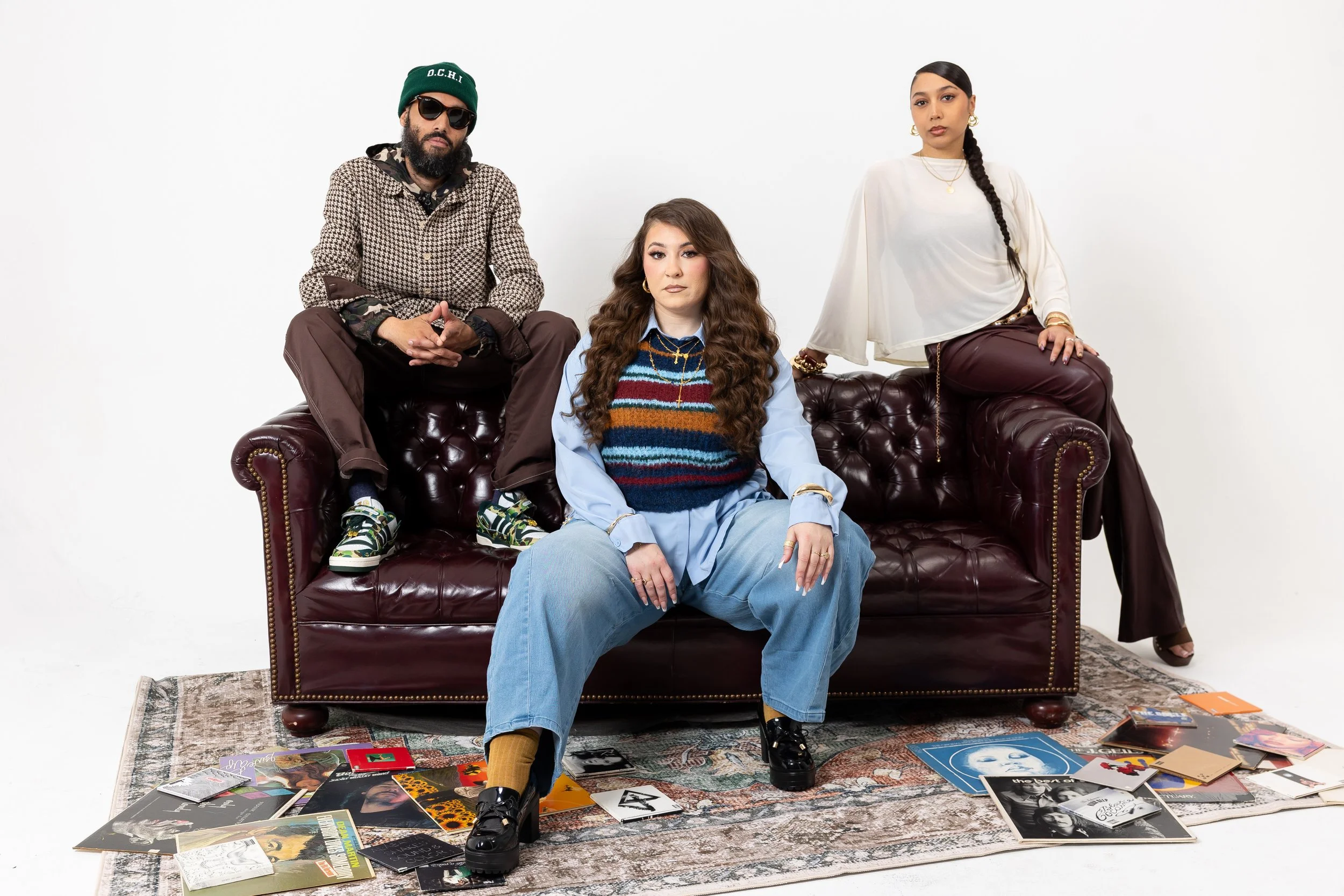 Three women sitting on a leather couch with vinyl records, magazines, and photo albums scattered on the floor in front of them against a white background.