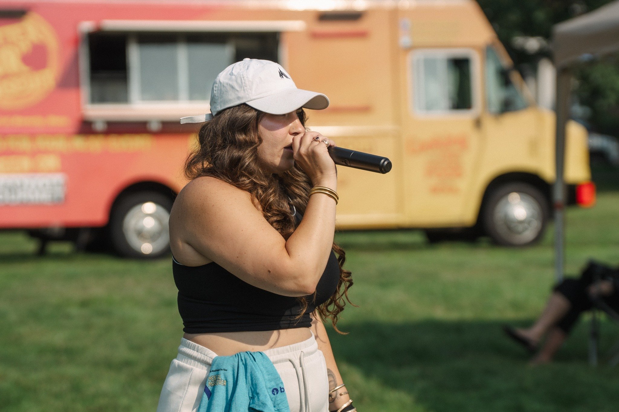 A young woman with long curly hair wearing a white baseball cap, black tank top, and white shorts, holding a microphone in an outdoor setting with a food truck in the background.