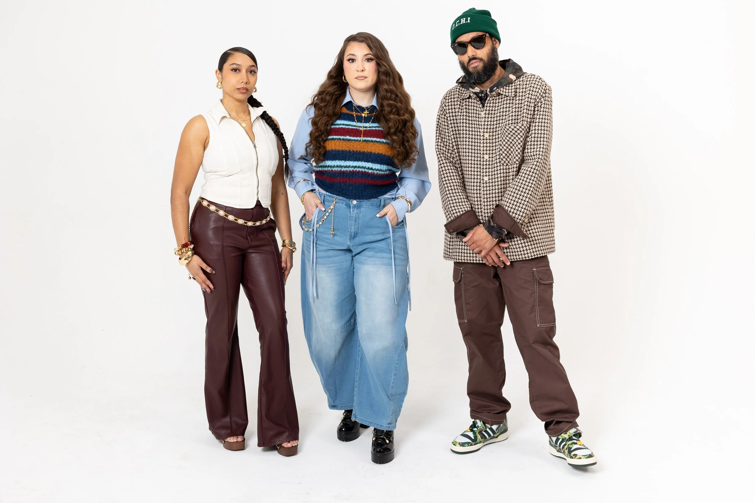 Three diverse people standing against a plain white background. The woman on the left has dark hair in a braid, wearing a white sleeveless top and maroon leather pants with accessories. The woman in the middle has long curly hair, wearing a multicolored striped sweater vest, blue shirt, and baggy jeans. The man on the right has a beard, sunglasses, a green beanie, and wears a patterned jacket with camouflage sneakers.