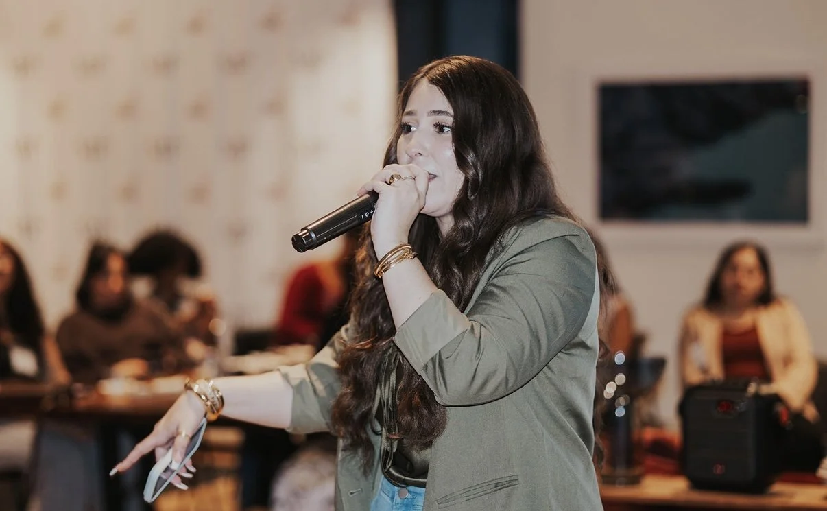 Young woman speaking into a microphone at a conference or meeting, with several people seated in the background.