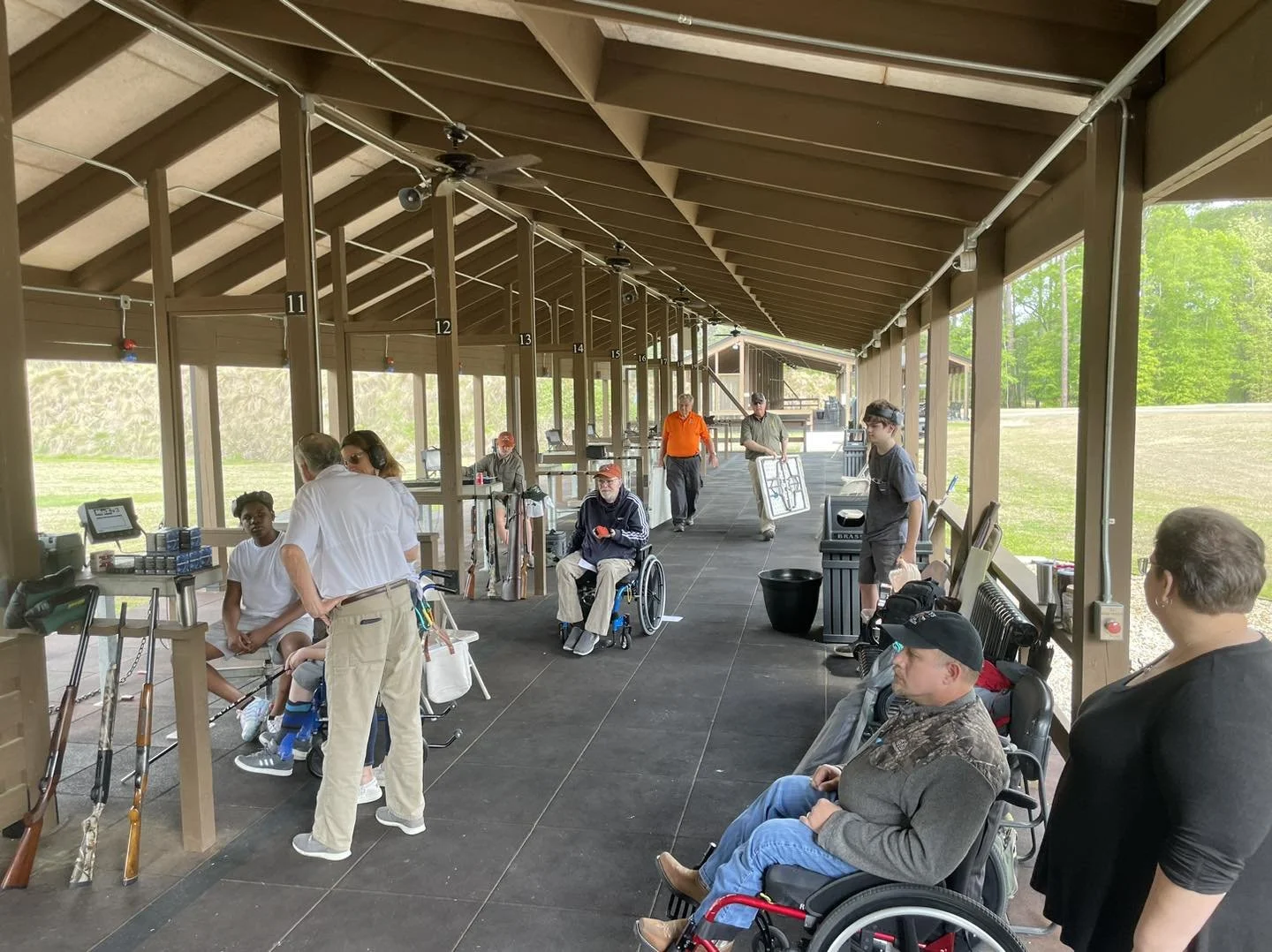 People are at a shooting range, some seated, some standing, with targets, firearms, and shooting paraphernalia visible. The structure is a covered outdoor shooting range with wooden beams and partitioned lanes.