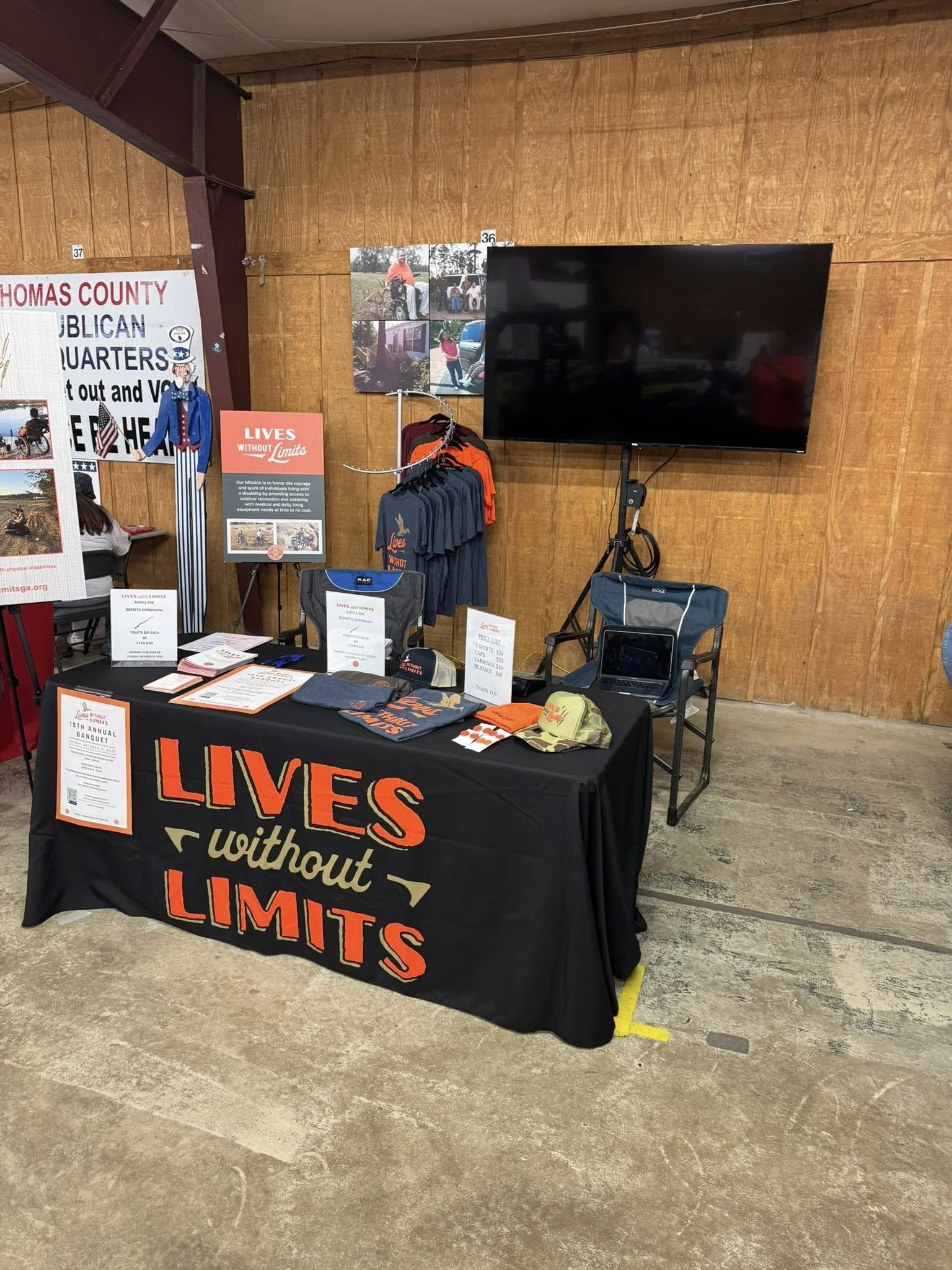Booth with a black tablecloth that reads "Lives Without Limits" and informational materials, clothing, and hats on display, against a wooden wall with a large TV screen and posters.