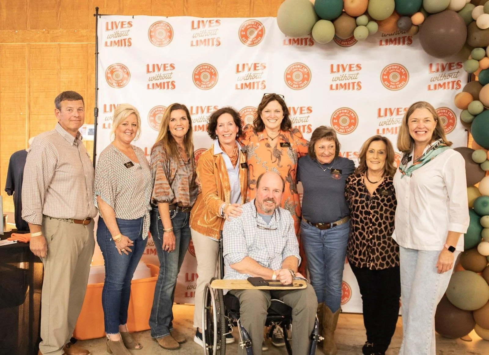 Group of nine people, including one in a wheelchair, standing together at a support event with a backdrop reading 'LIVES without LIMITS' and a balloon arrangement on the side.