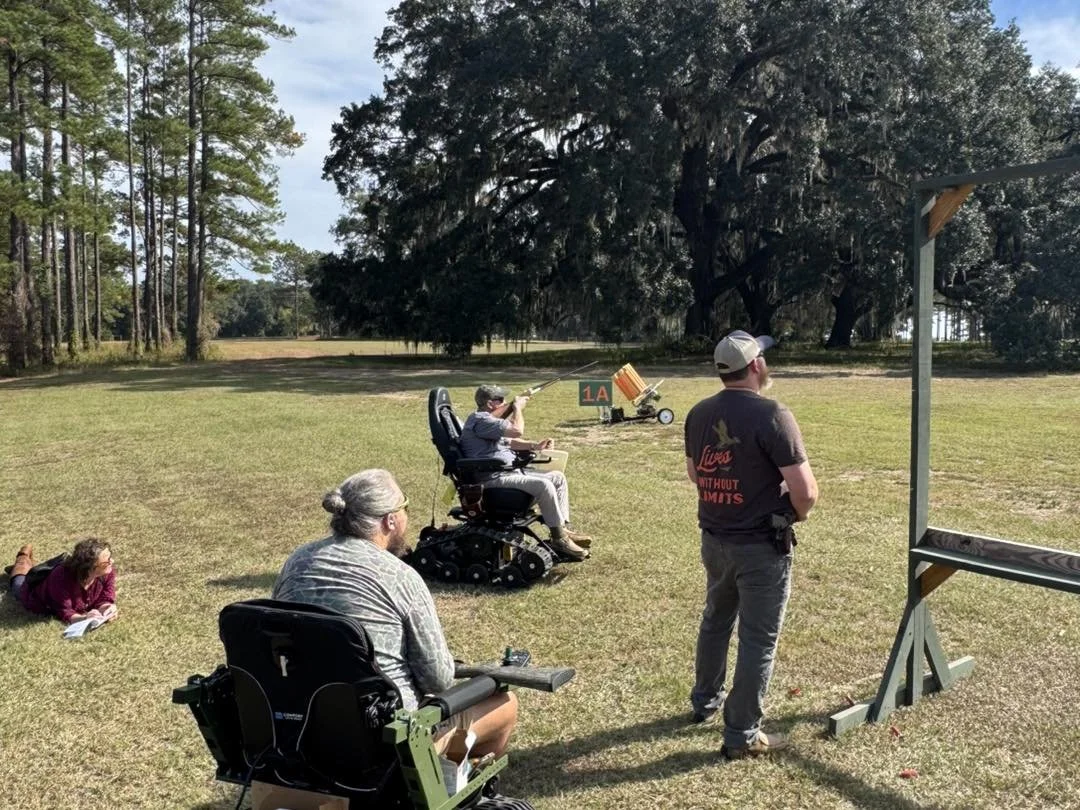 People participating in a shooting or archery activity outdoors on a grassy field with trees in the background. One person is aiming a rifle at a target, while others are seated or lying on the ground nearby.